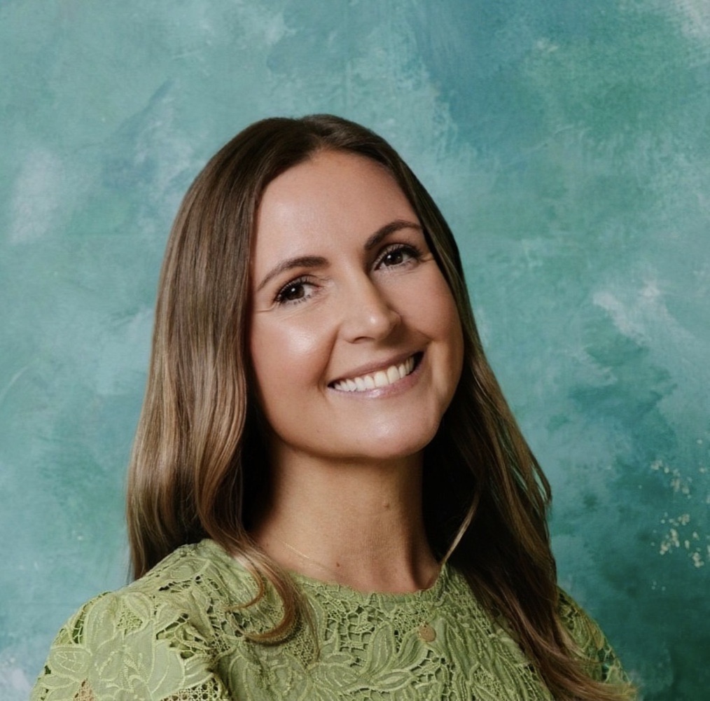 Smiling woman with long brown hair wearing a green lace top, against a textured teal background.