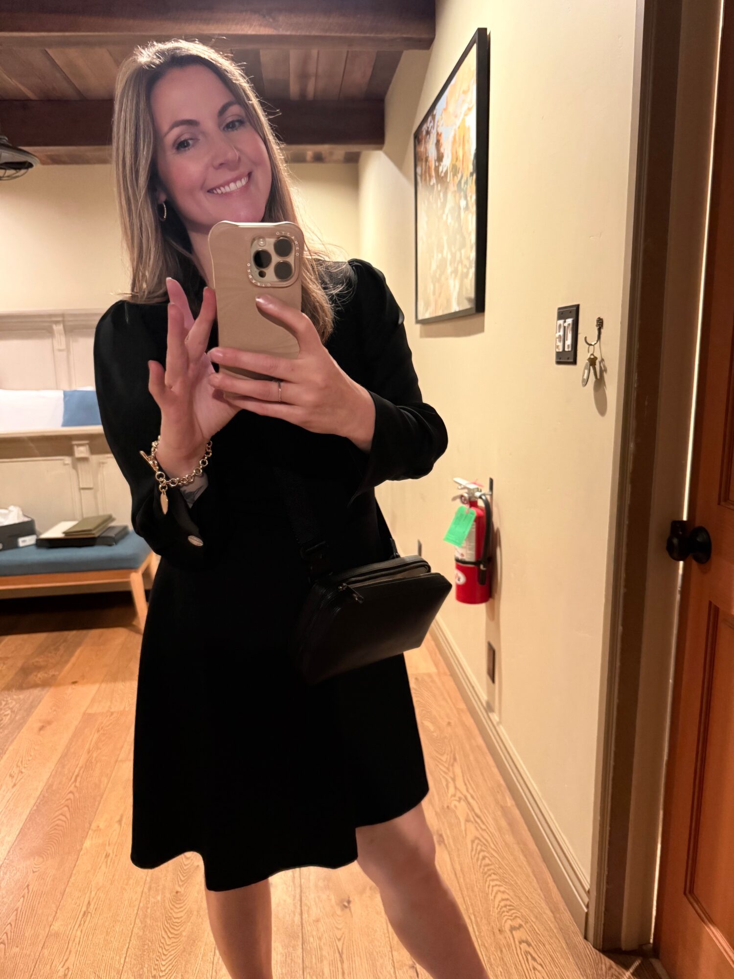 Woman taking a mirror selfie, smiling, in a black dress, holding a phone, in a room with wooden floors and artwork on the wall.