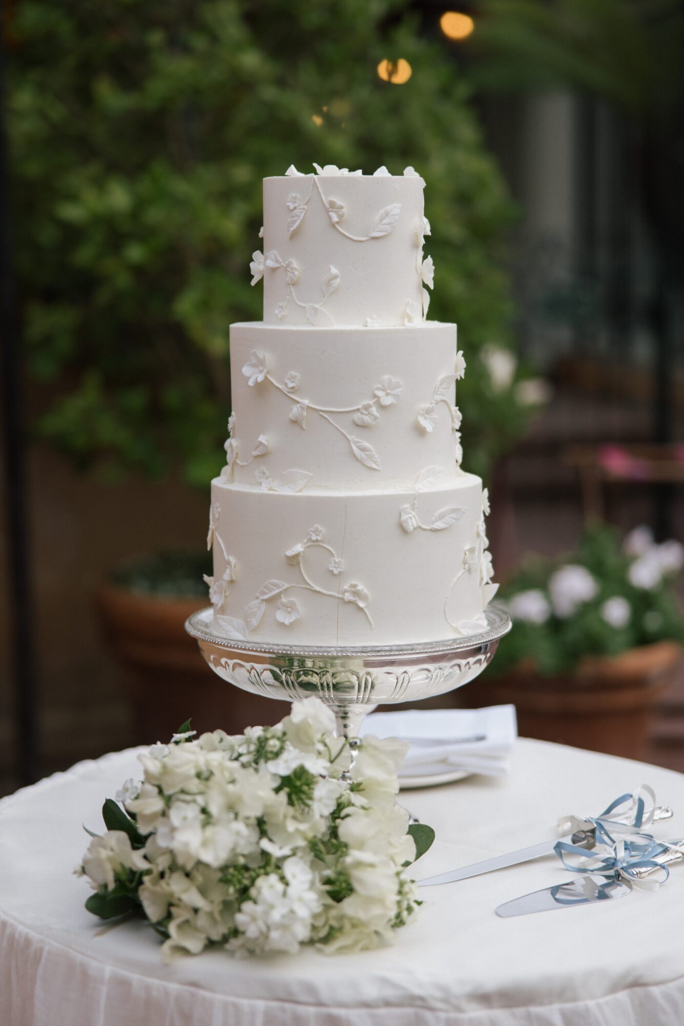 Three-tiered white wedding cake with floral decorations on a glass stand, with a bouquet of white flowers and ribbons on table