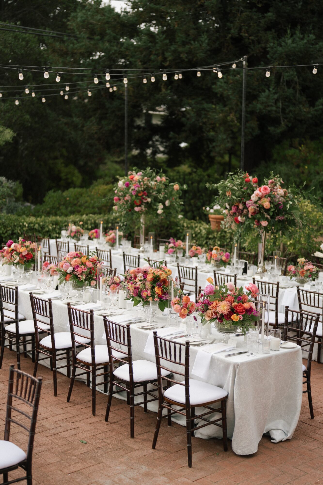 Outdoor event setup with long tables, floral centerpieces, string lights, and chairs on a brick surface, surrounded by trees.
