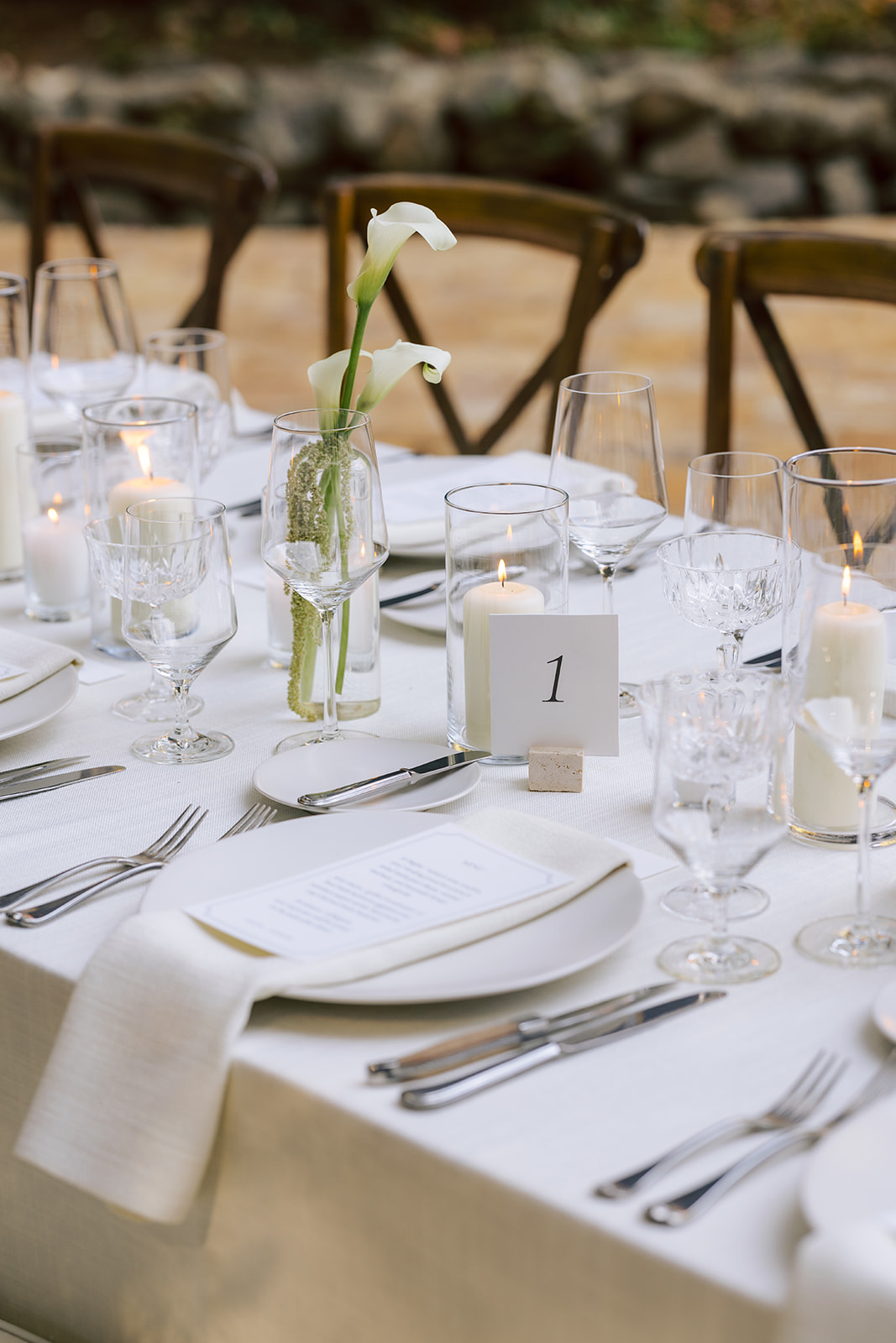 Table set with white tablecloth, glassware, candles, and a small flower arrangement, outdoors with chairs in background.