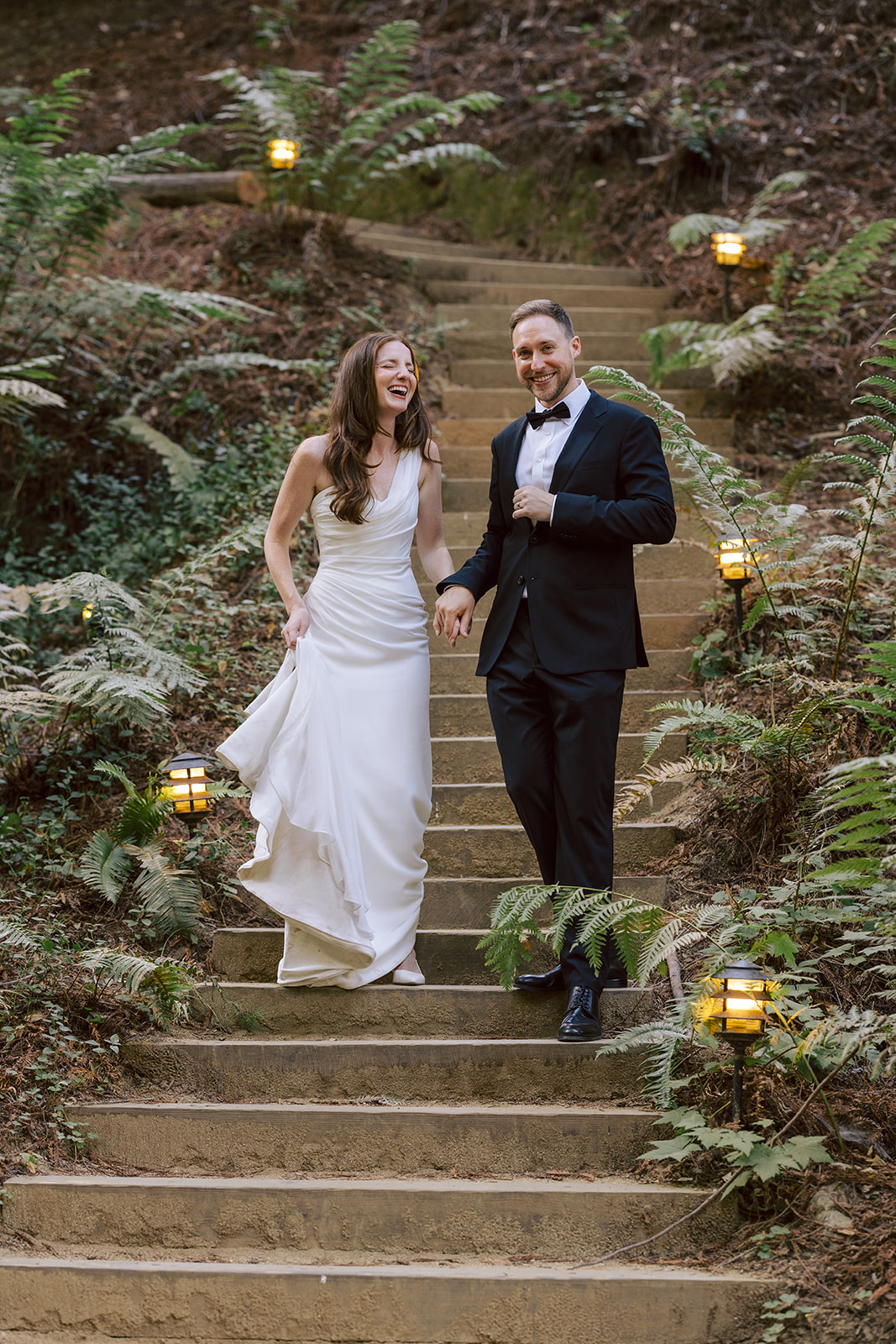 A bride and groom walking down outdoor stone steps surrounded by greenery and lanterns, smiling and holding hands.