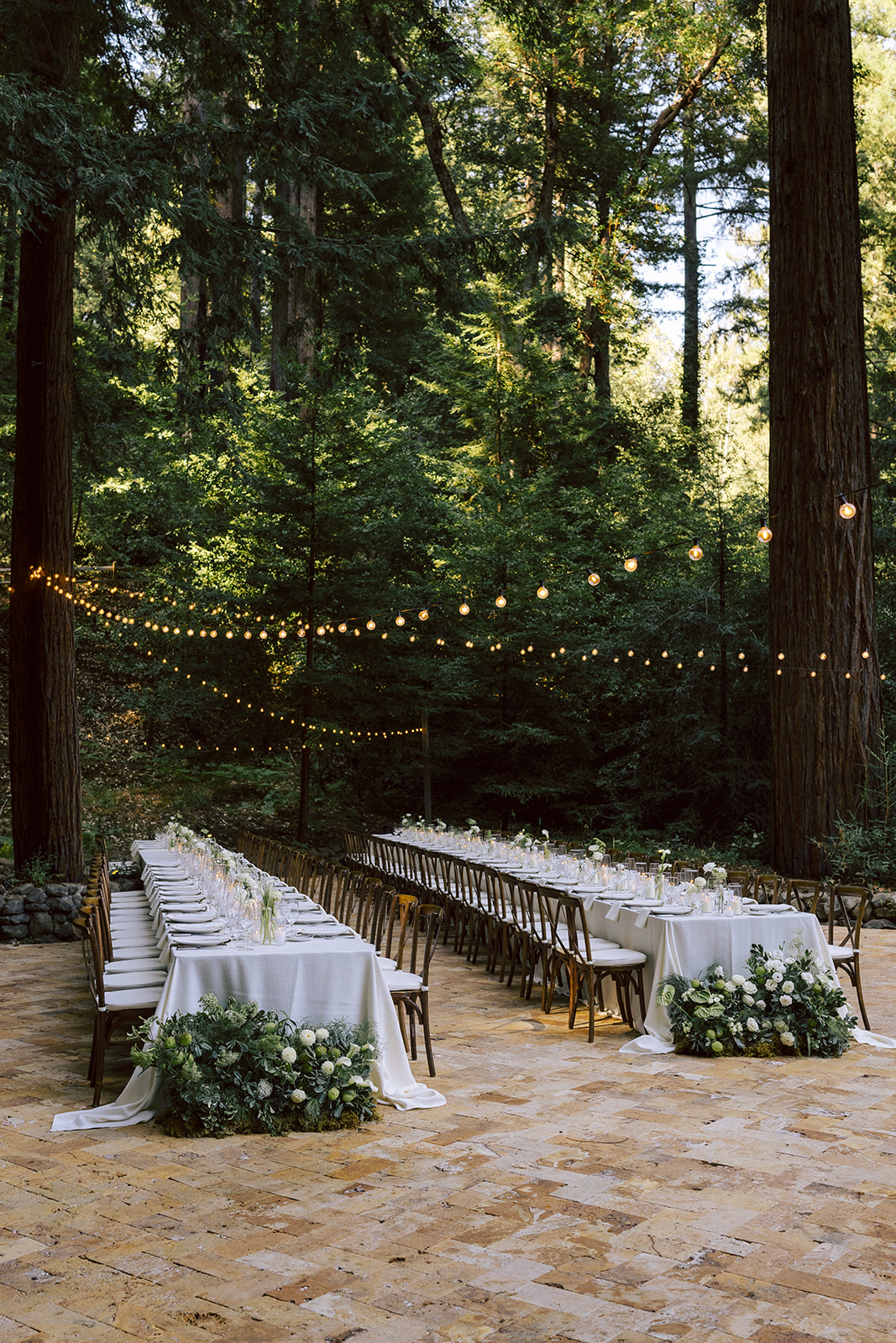 Outdoor event setup with long tables, white tablecloths, floral arrangements, and string lights in a forest clearing.