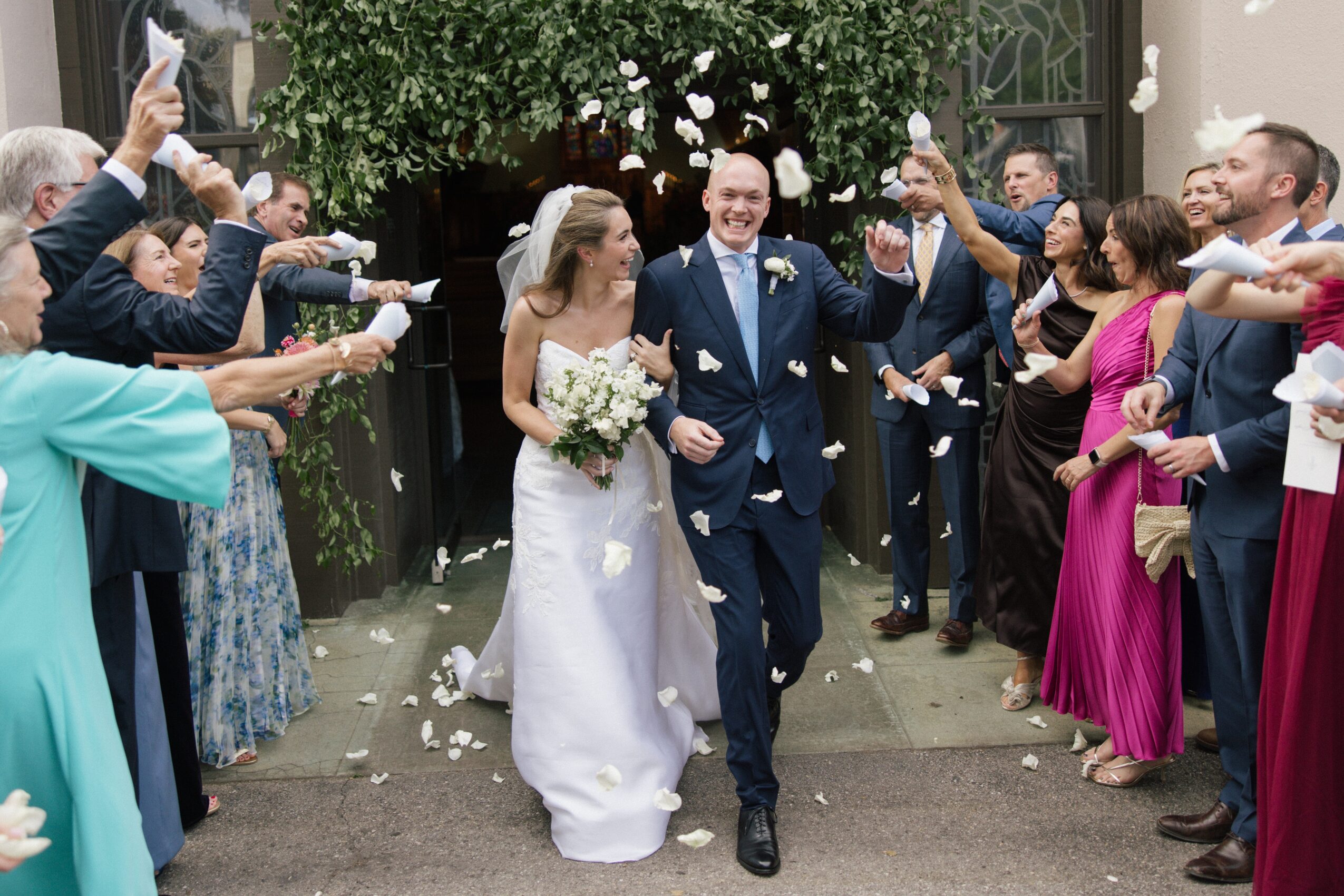 Bride and groom walking together surrounded by guests throwing confetti, outdoors with greenery and flowers, celebrating wedding.