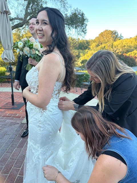 Woman in wedding dress smiling outdoors, with two women adjusting her dress, trees and sky in background.