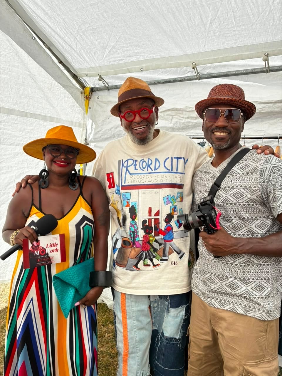 Three people standing together inside a tent, smiling, wearing colorful clothing and hats, with one holding a camera.