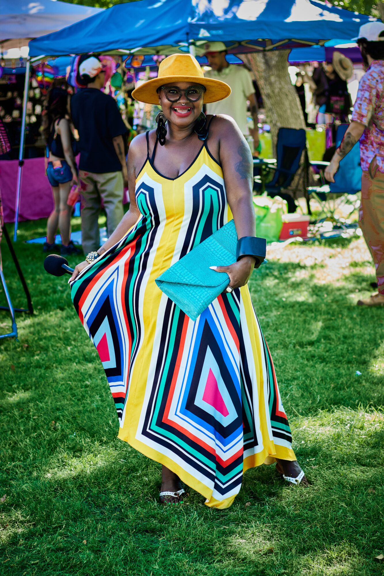Woman in a colorful striped dress and yellow hat holding a blue clutch at an outdoor market, smiling.
