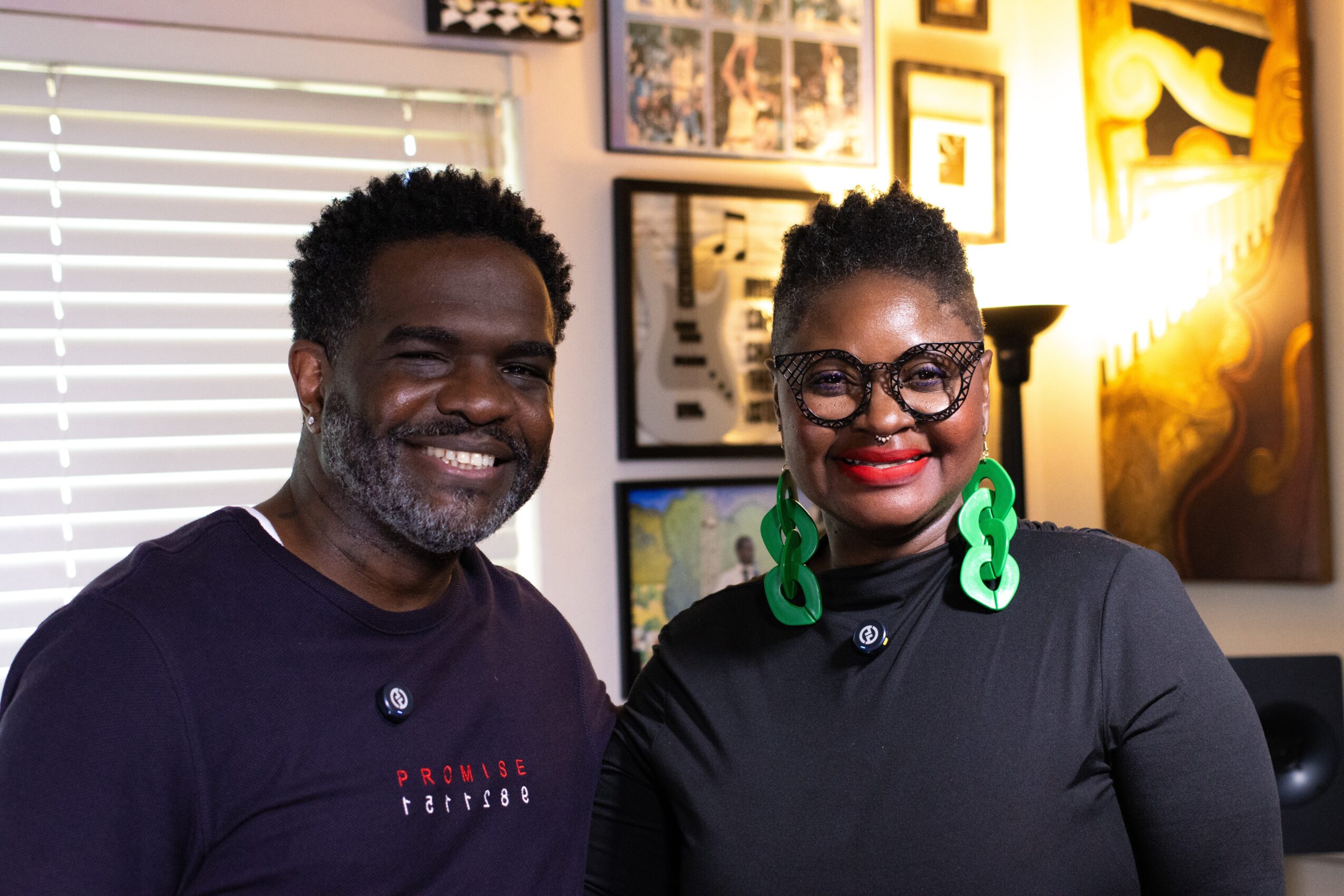 Two people smiling indoors, one with glasses and large green earrings, in front of framed artwork and a window with blinds.