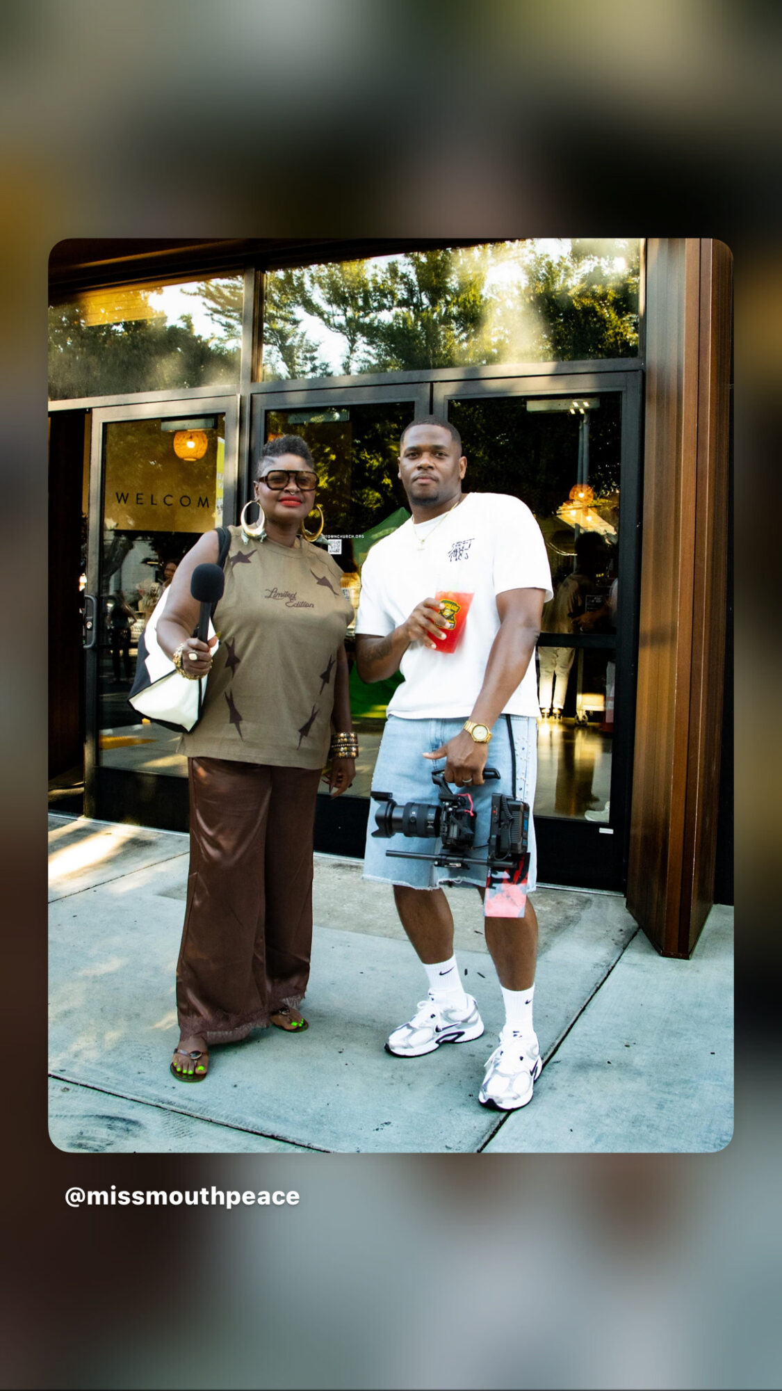 Two people standing outside a building, one holding a drink, smiling, with trees and glass doors in the background.
