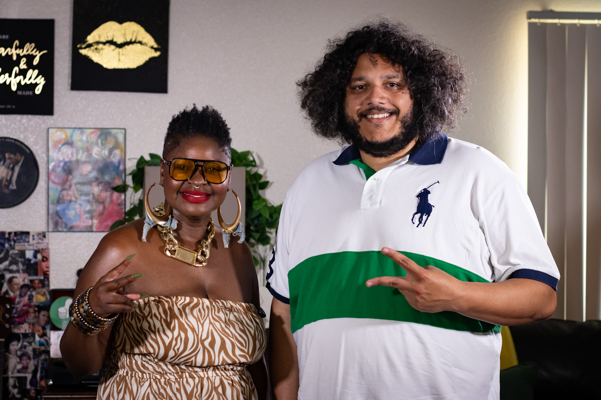 Two people smiling indoors, one with curly hair and the other with short hair and sunglasses, standing in front of a wall with pictures.