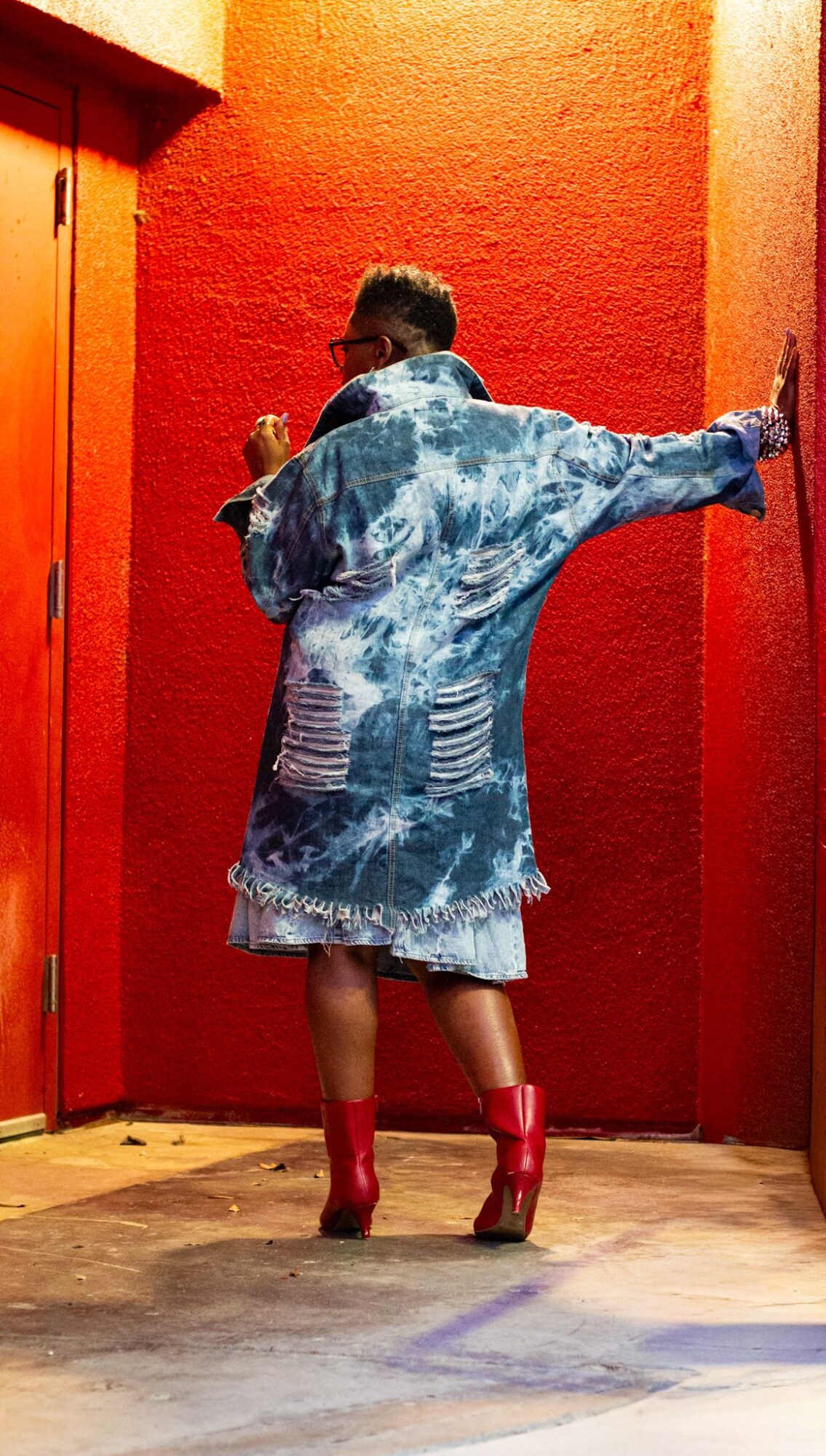Person standing with back to camera, wearing a denim jacket, shorts, red boots, against a red wall.