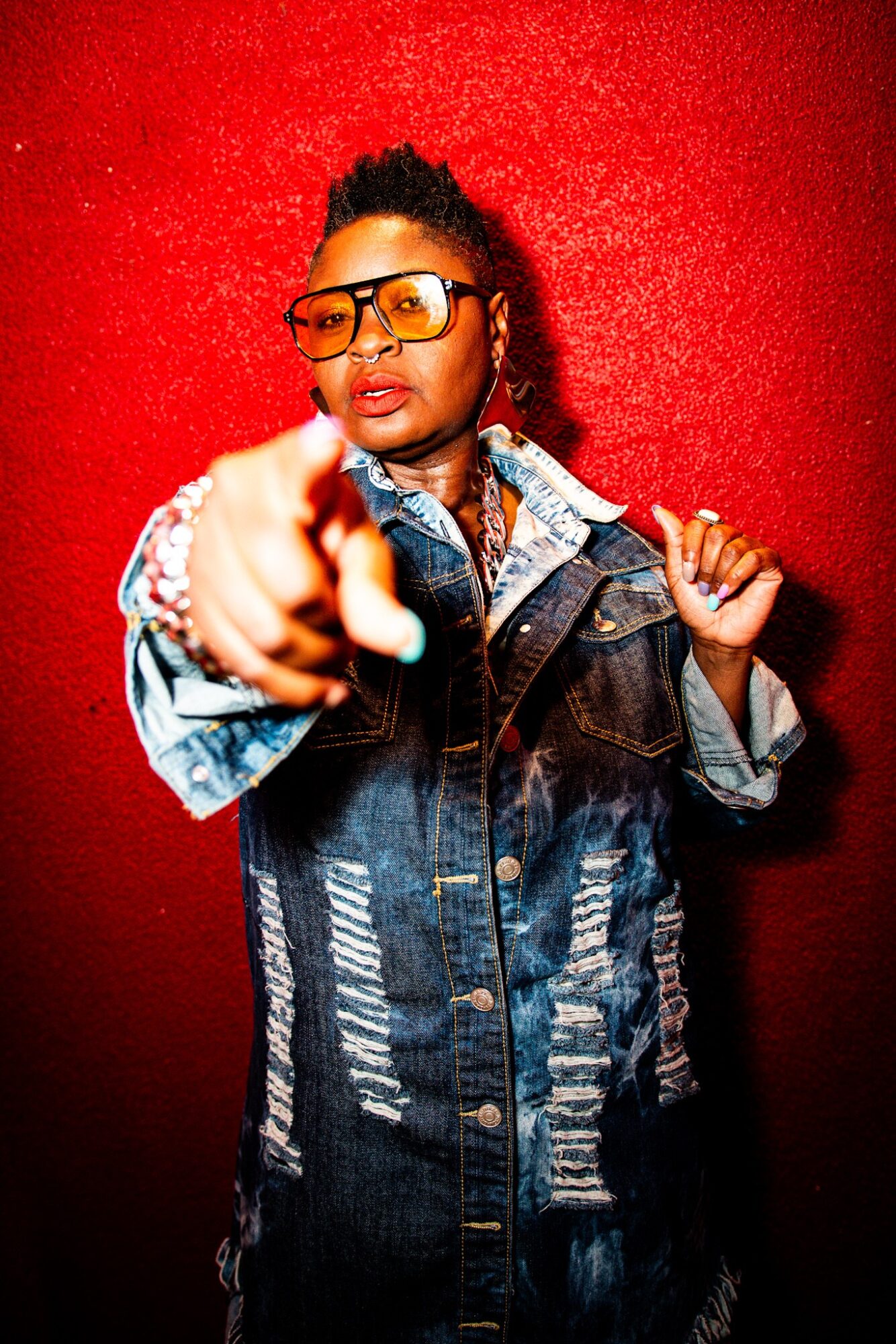 Person wearing glasses and denim jacket pointing forward against red background.