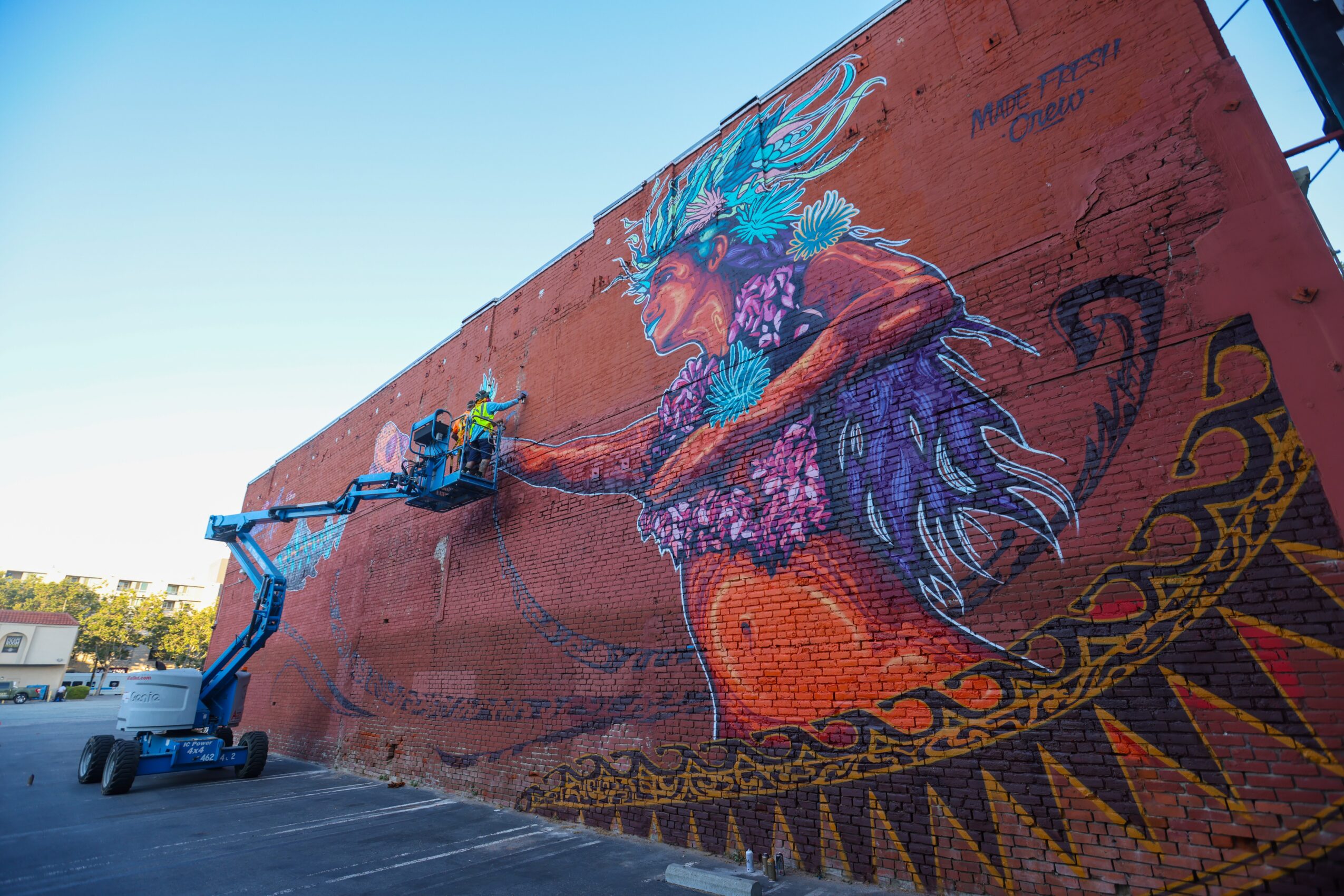 Mural of a colorful mythical creature with a long, flowing tail on a brick wall, with a person working on it using a lift.