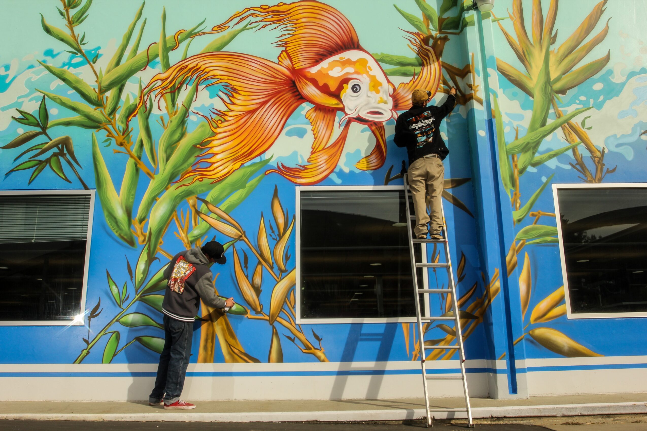 Two people painting a colorful mural of a fish and aquatic plants on a wall with windows.