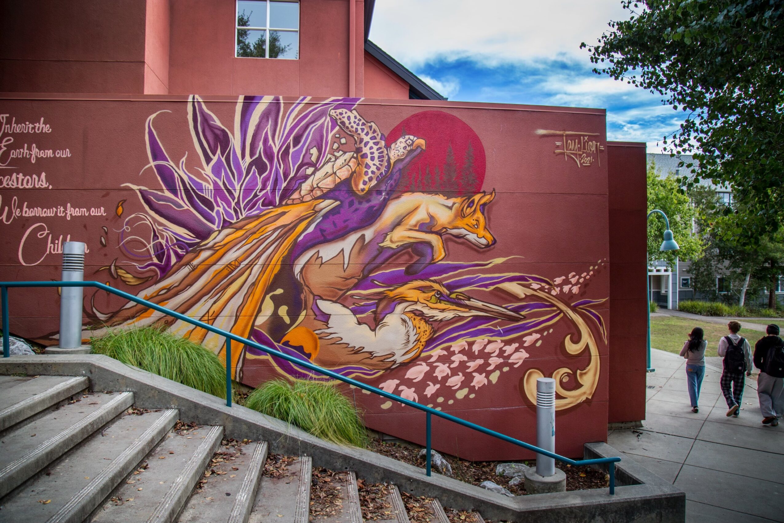 Colorful mural of a peacock with vibrant feathers on a building wall, with trees and people nearby.