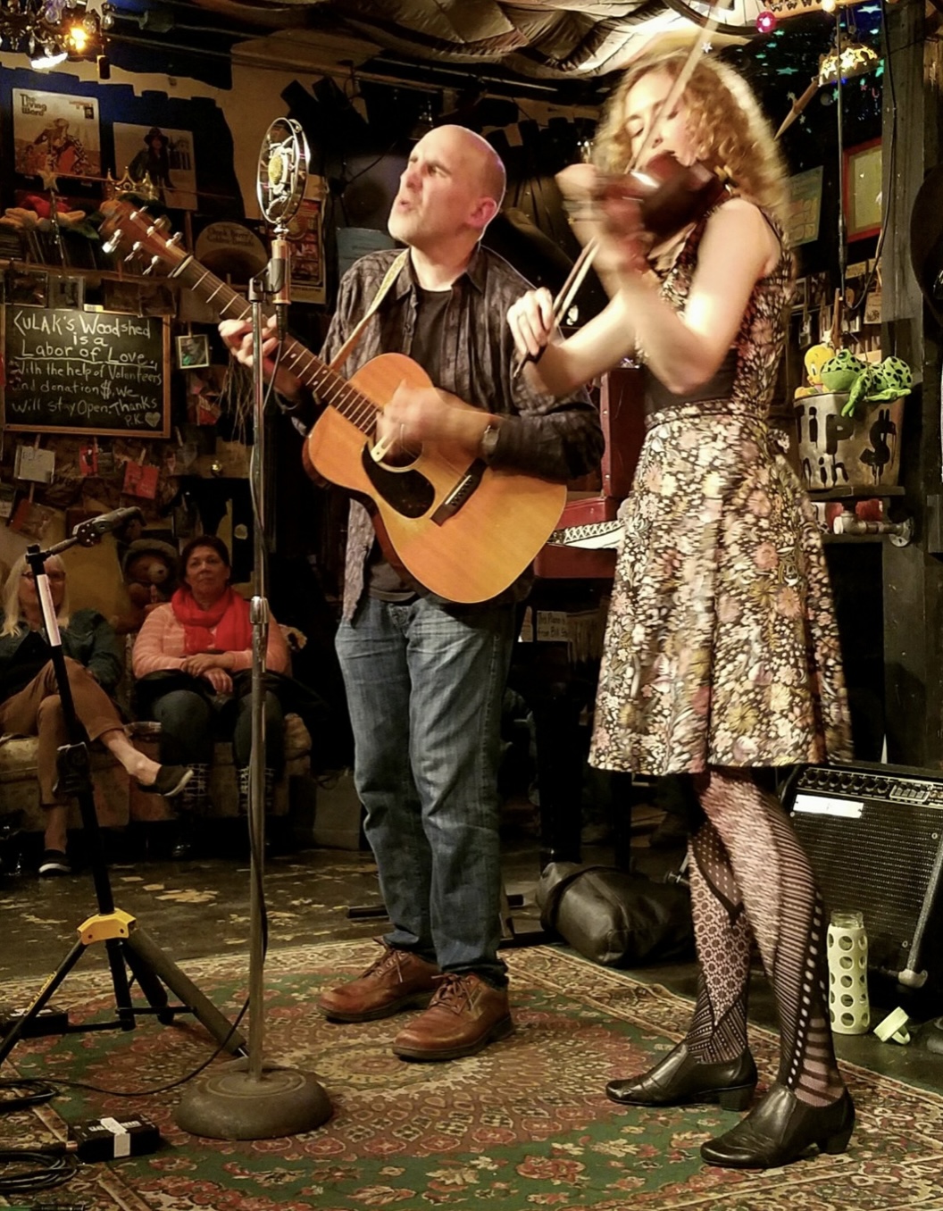 Man playing guitar and woman singing or speaking on stage in a lively venue with audience and decorations.