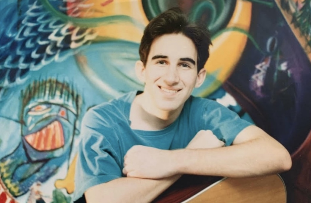 Young man with dark hair smiling, leaning on a table with colorful graffiti background