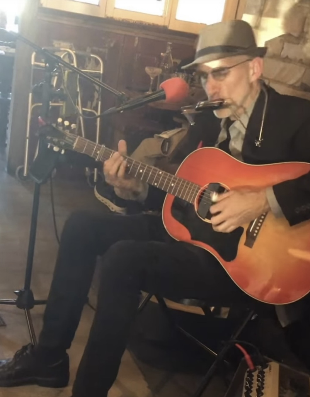 Person wearing a hat and sunglasses playing an acoustic guitar with a microphone nearby, seated indoors.