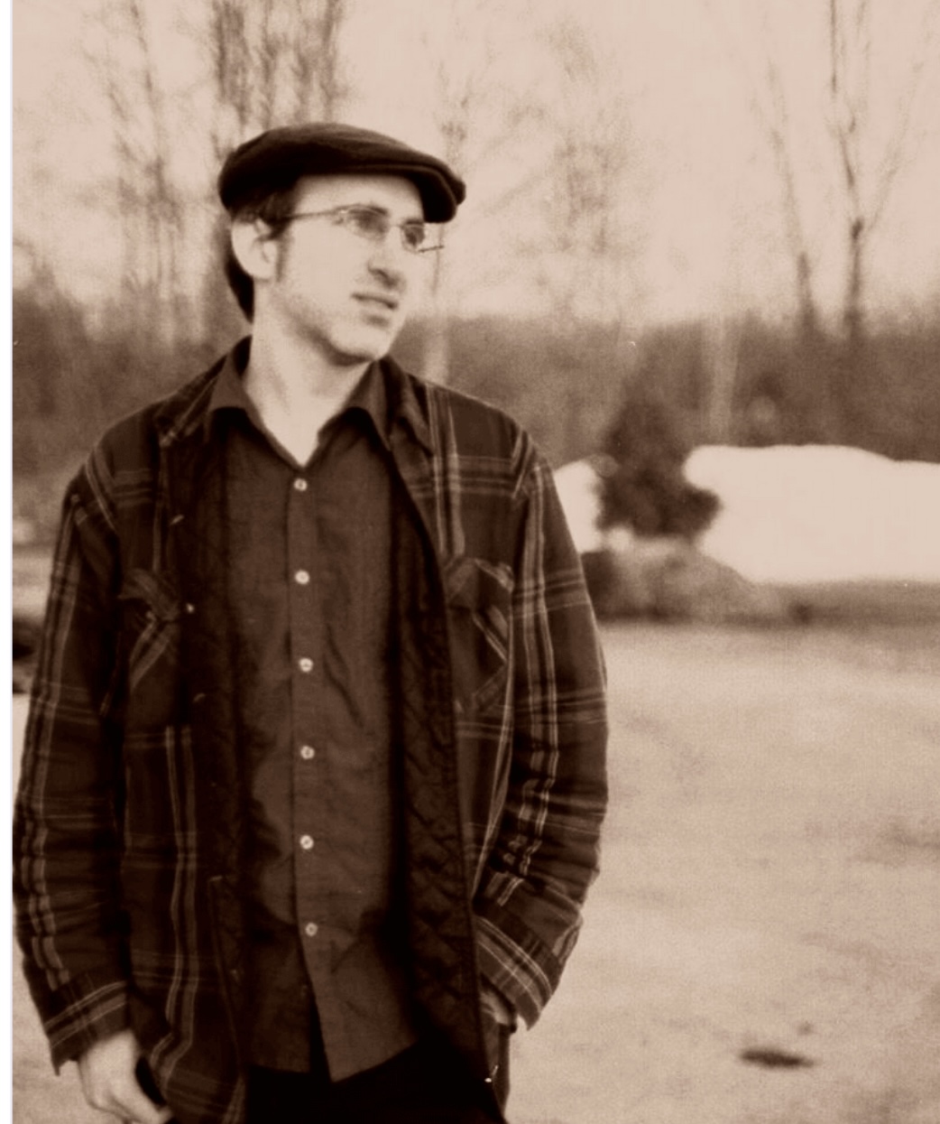 Young man with glasses and dark hair, wearing a plaid jacket and shirt, standing outdoors with trees in background.