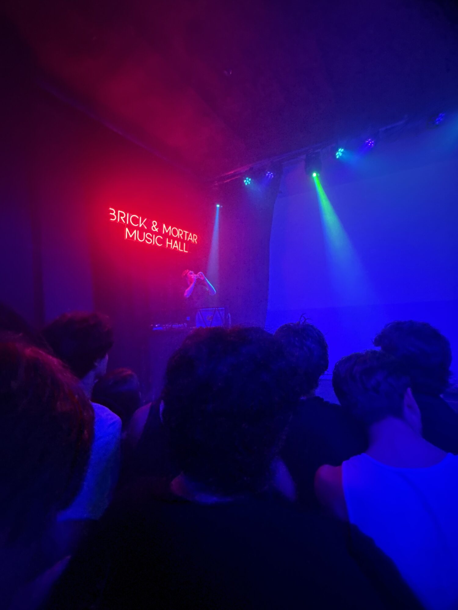People in a dark room with colorful lights and a red sign that reads 'BRICK & MORTAR MUSIC HALL'.