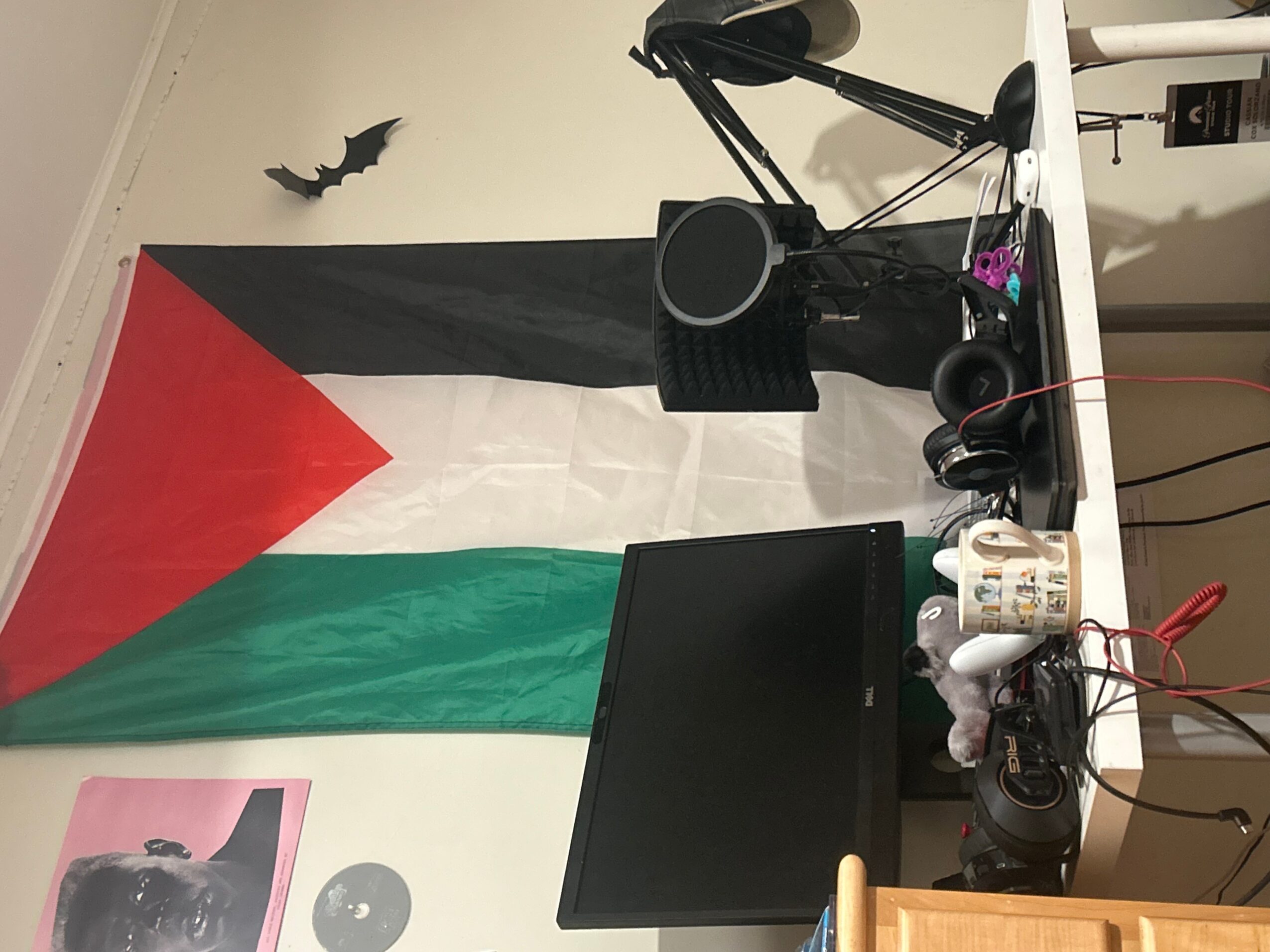 Palestinian flag hanging on wall with a small bat decoration, desk with monitor, headphones, and various items, and a white wall.