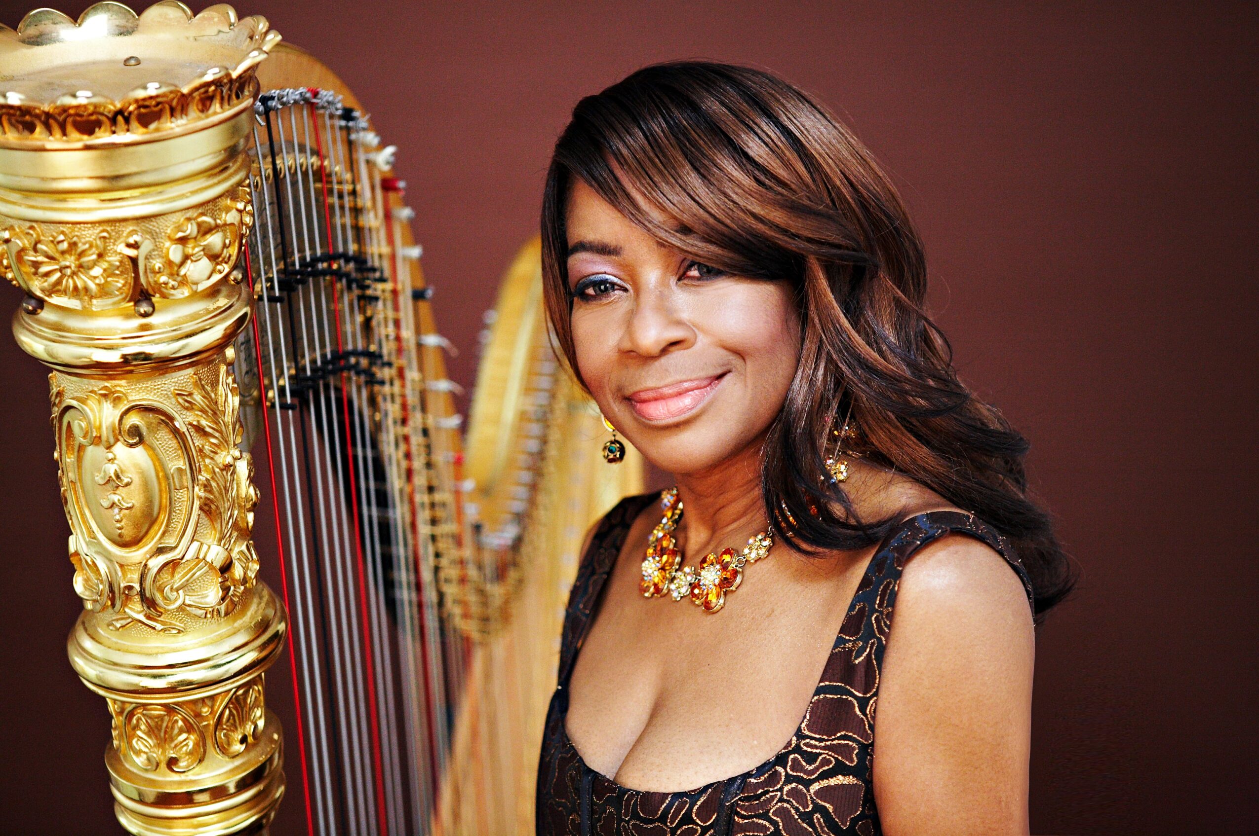 Woman with dark hair and a necklace, standing next to a golden harp, smiling, against a brown background.