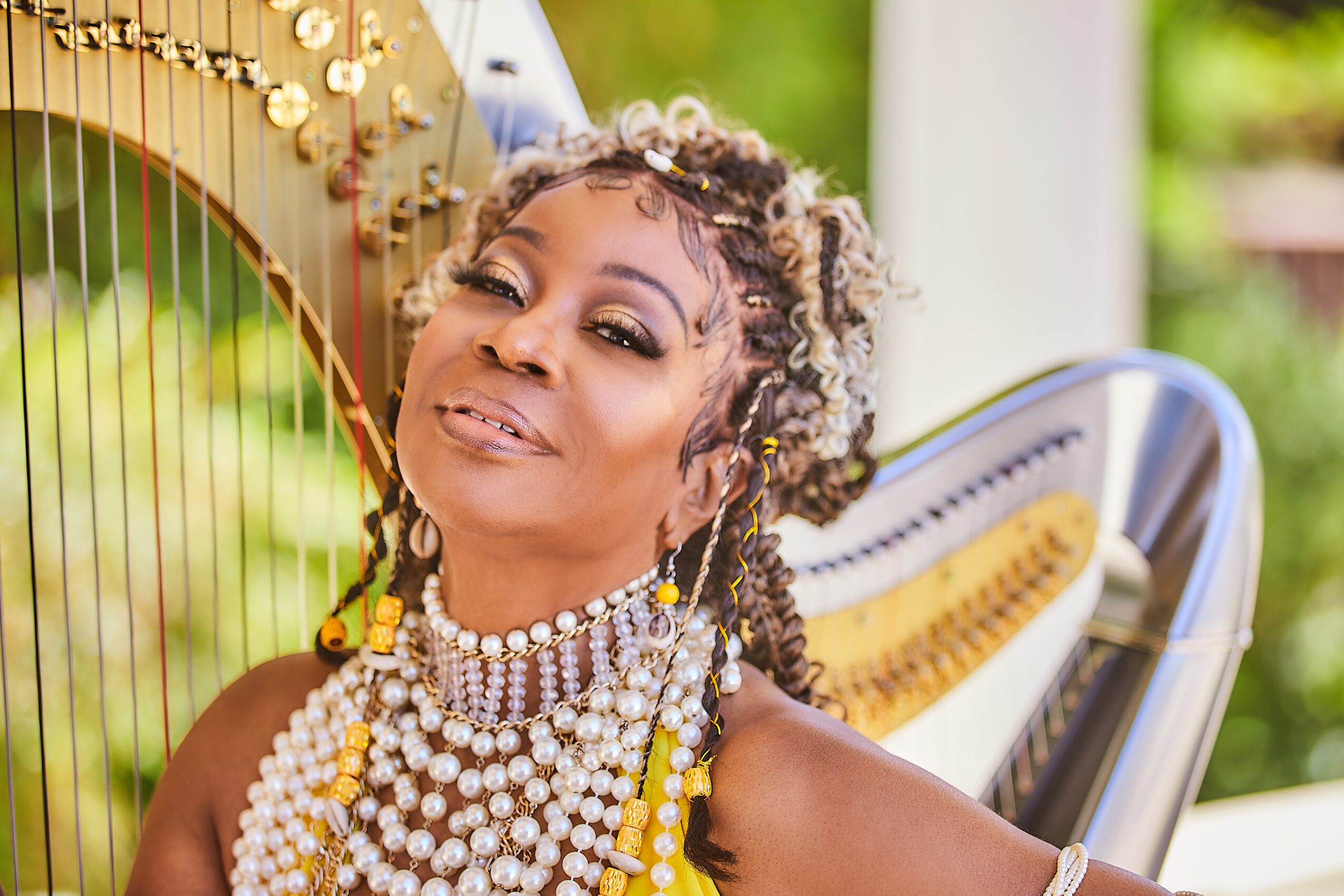 Woman with curly hair wearing multiple pearl necklaces, smiling outdoors near a harp, with greenery in background.