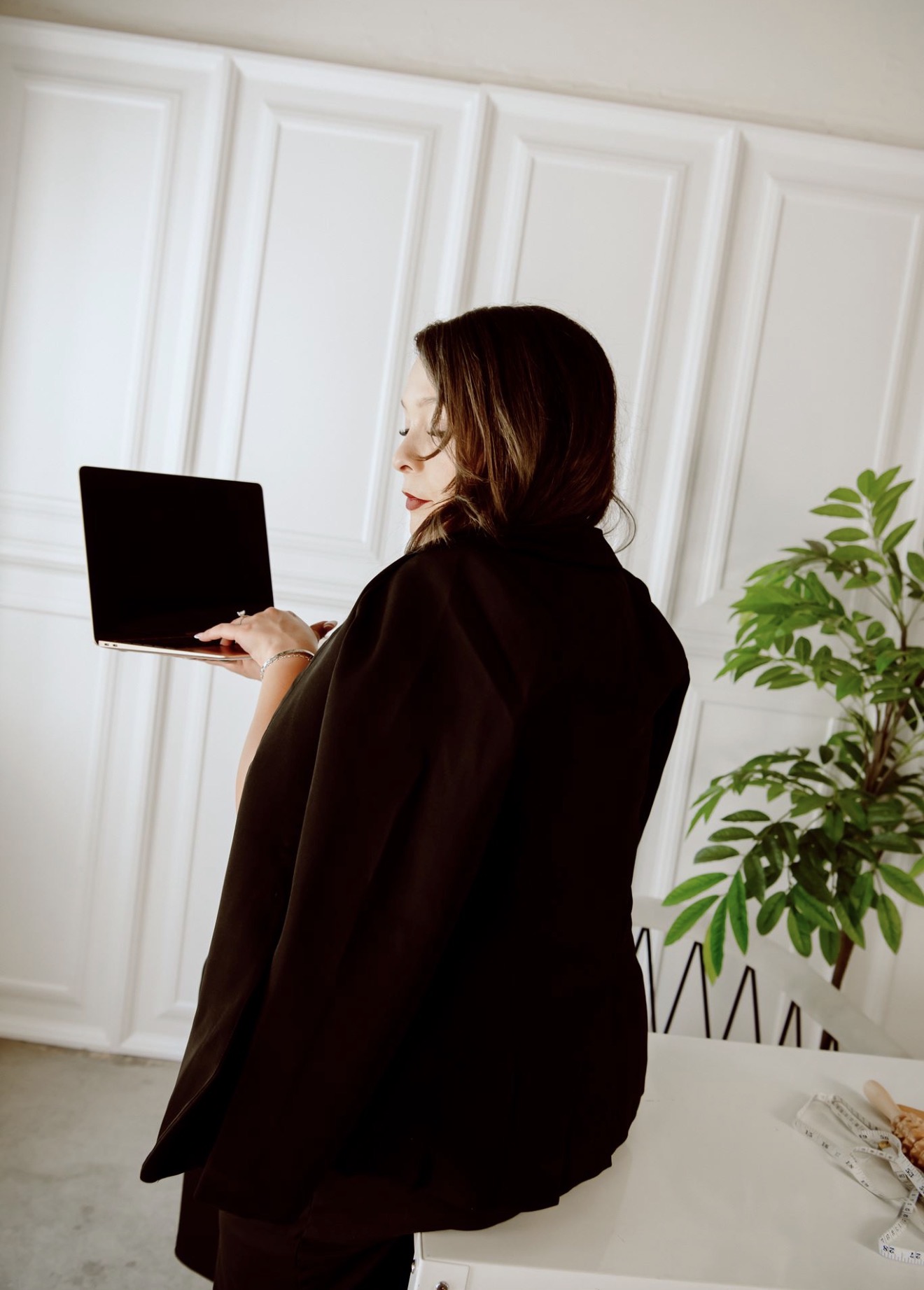 Woman with dark hair in a black coat holding a laptop, standing near a white wall and a green plant.