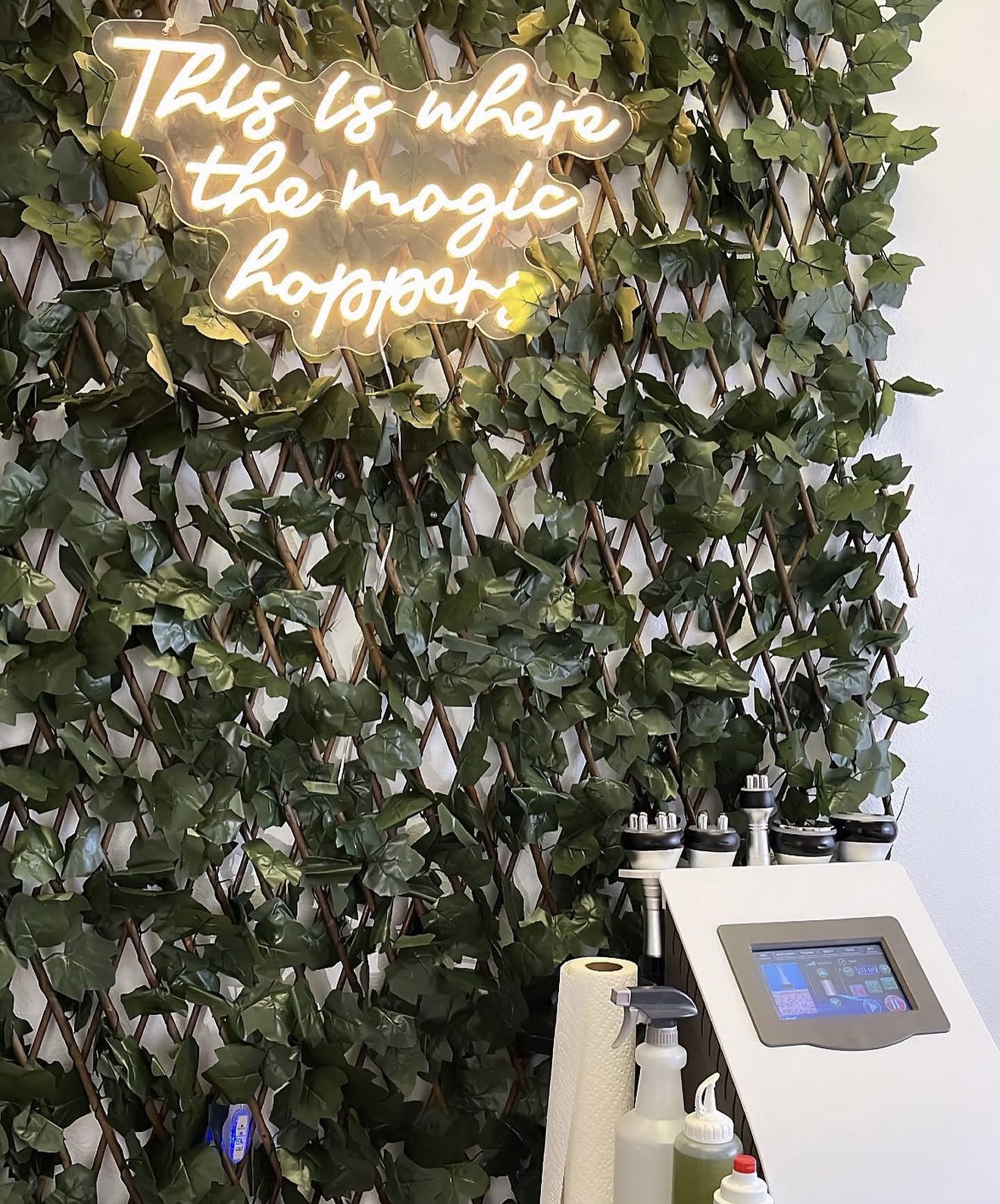 Neon sign reading 'This is where the magic happens' above a green leafy wall, with a white device and supplies below.