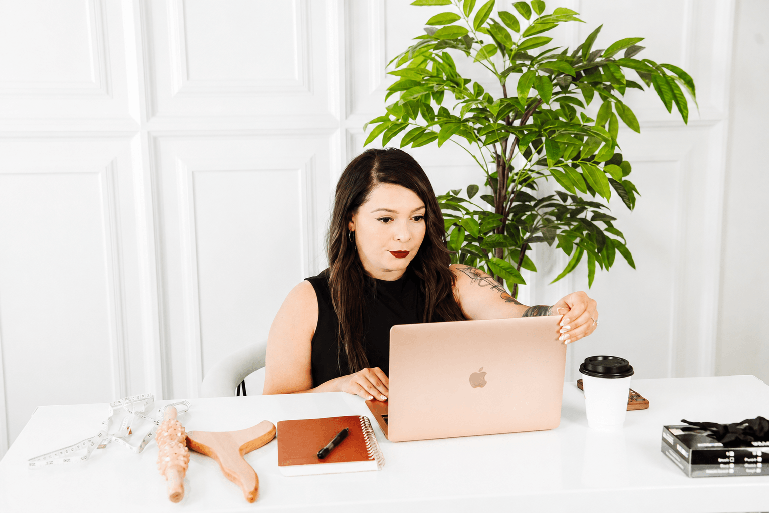 Woman with long dark hair working on a laptop at a white table, with a large green plant behind her.