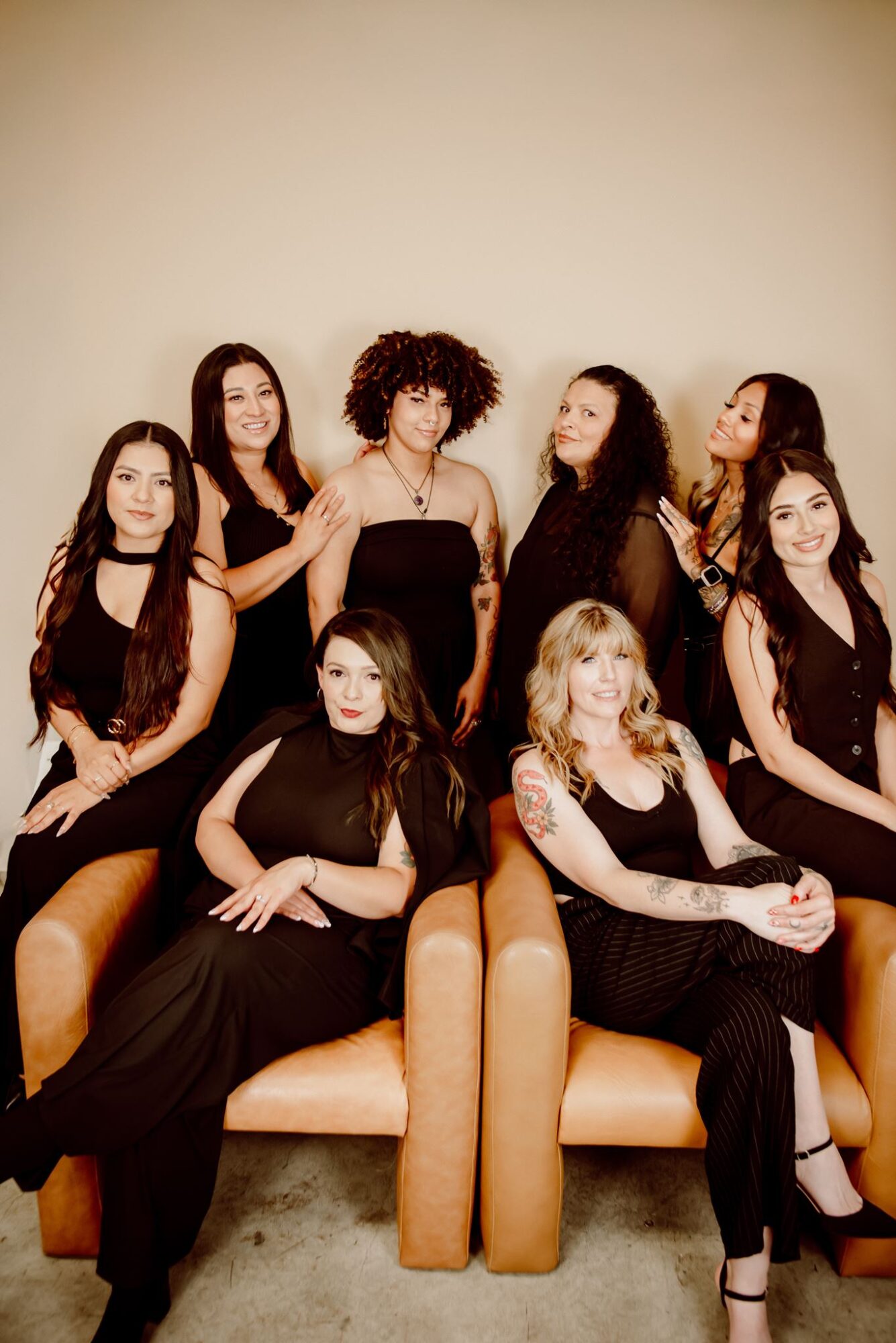 Seven women in black dresses sitting and standing around a beige sofa, posing for a photo against a plain background.