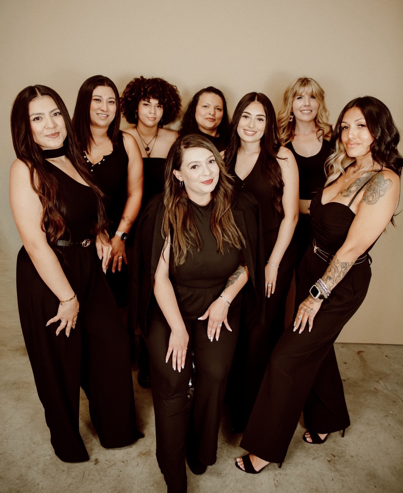 Group of eight women standing together against a plain wall, dressed in black outfits, smiling for the photo.