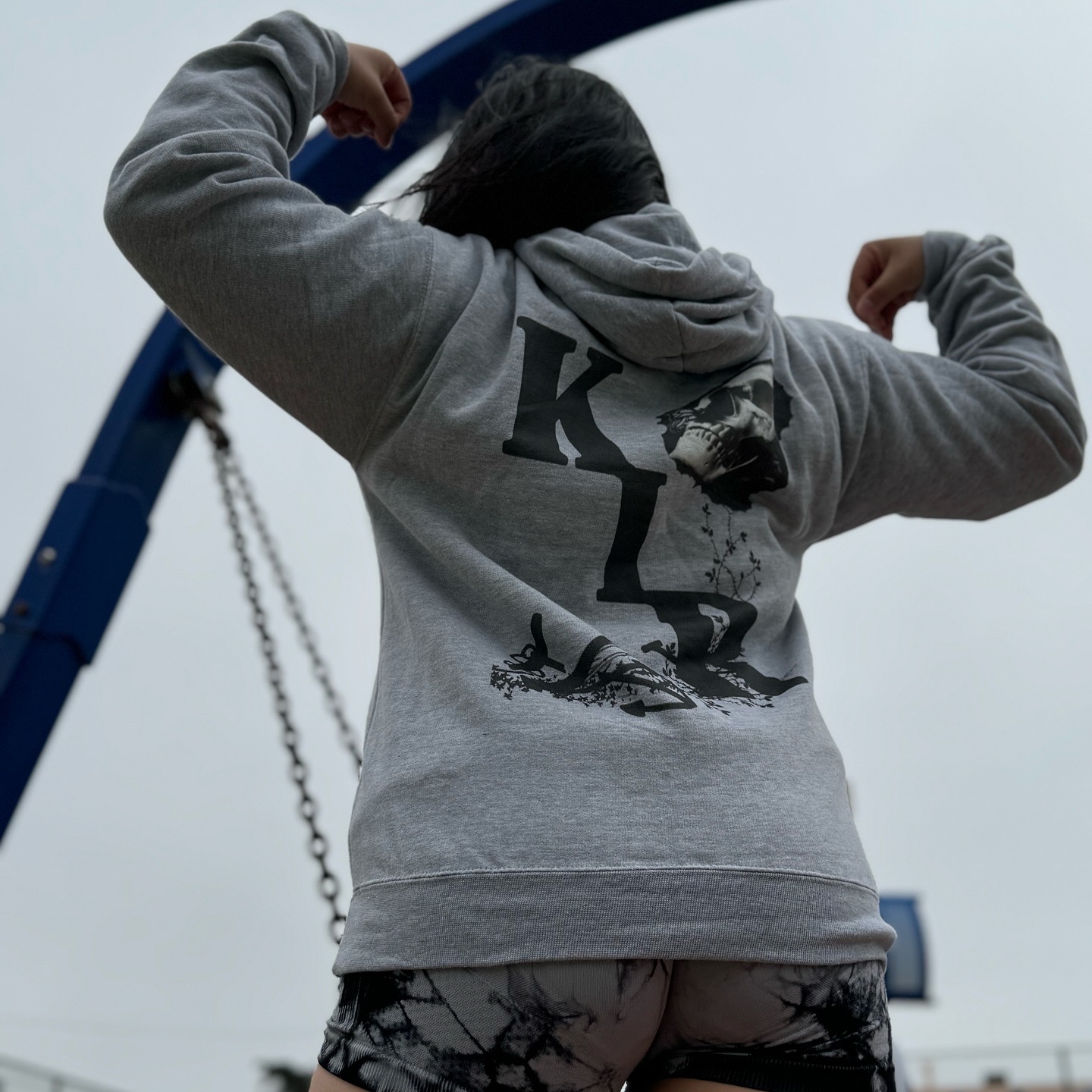 Person with dark hair wearing a gray hoodie and patterned shorts, flexing arms, with a swing set in the background.