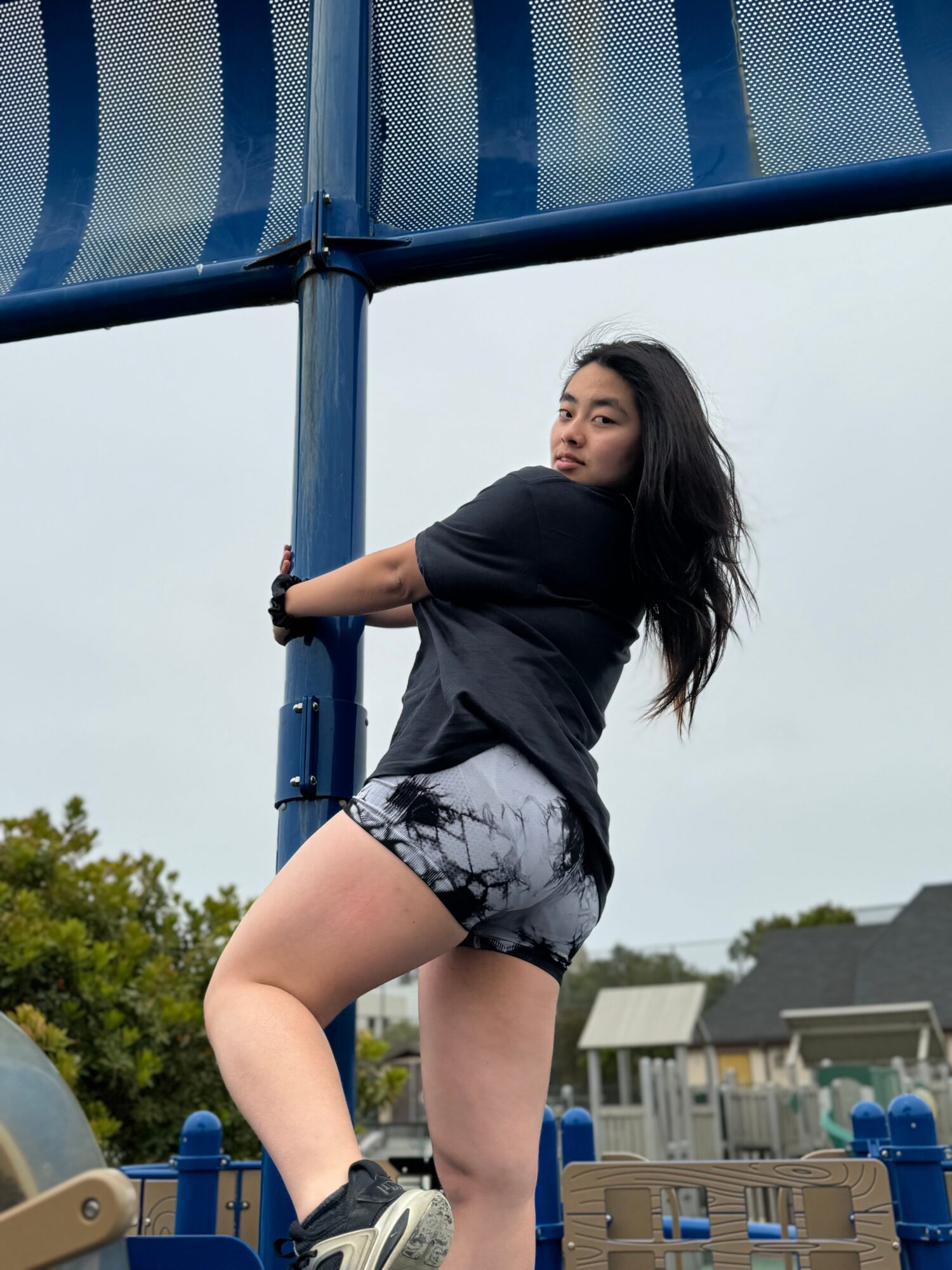 Young woman with long dark hair in black shirt and patterned shorts standing outdoors near playground equipment, looking over shoulder.