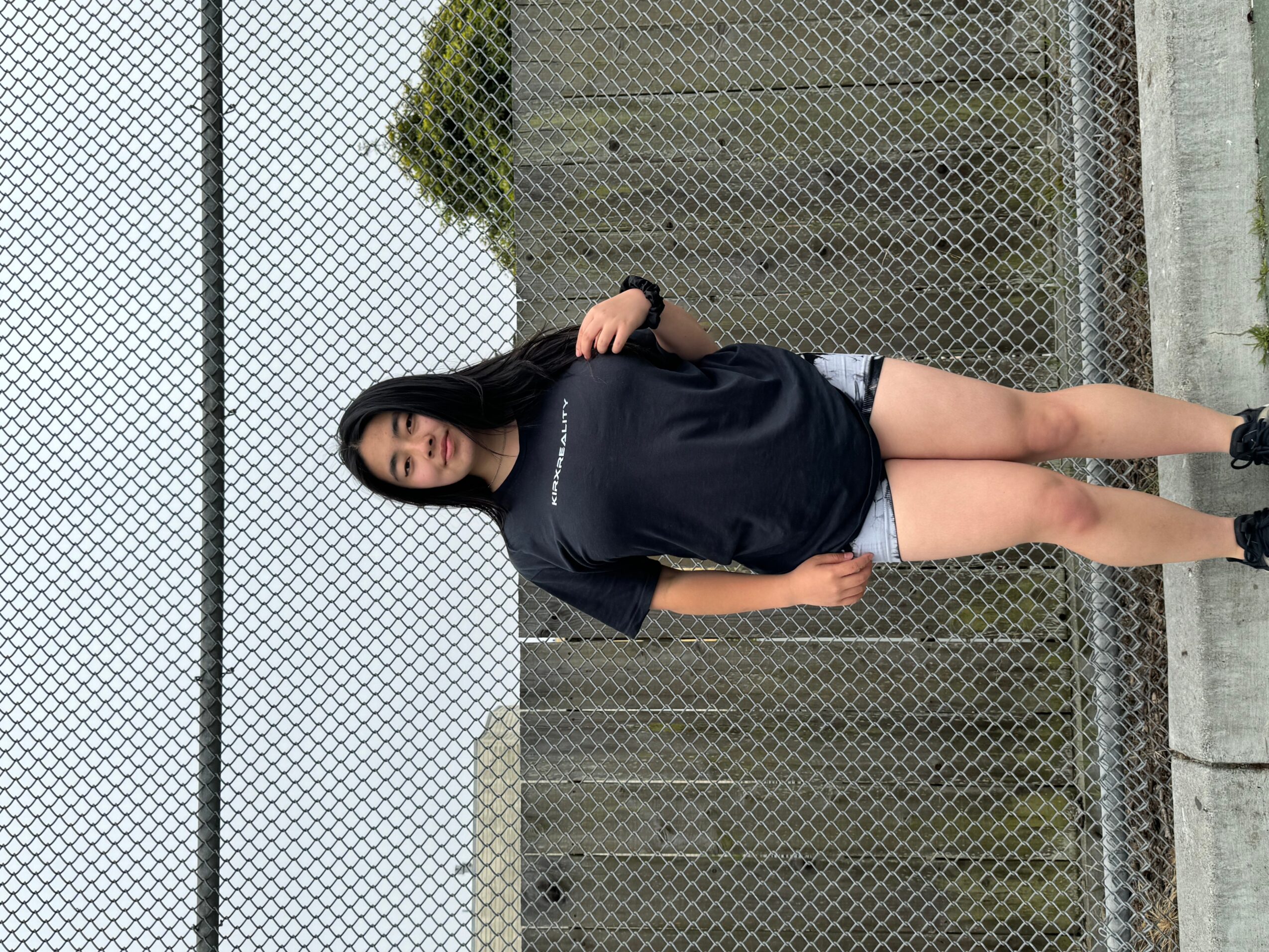 Young woman standing on a chain-link fence, wearing a black oversized t-shirt and shorts, with trees in the background.