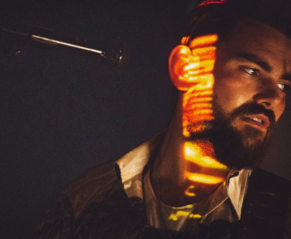 Portrait of a man with a beard and dark hair, illuminated by orange light patterns on his face and neck.