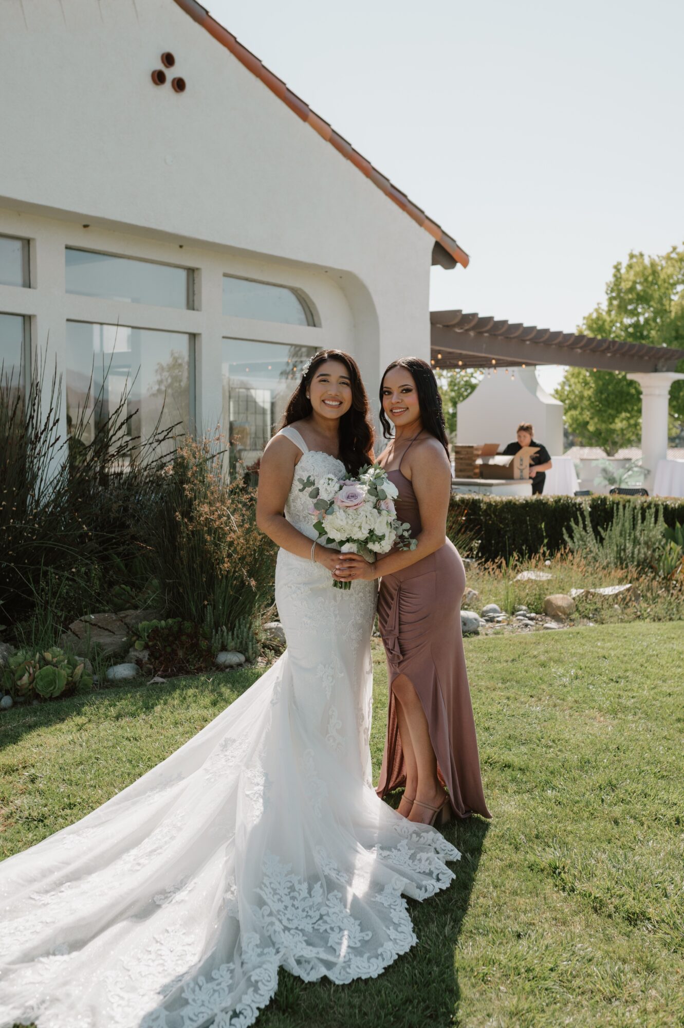 Two women in wedding and bridesmaid dresses standing outdoors, holding a bouquet, smiling, with a building and garden in background.