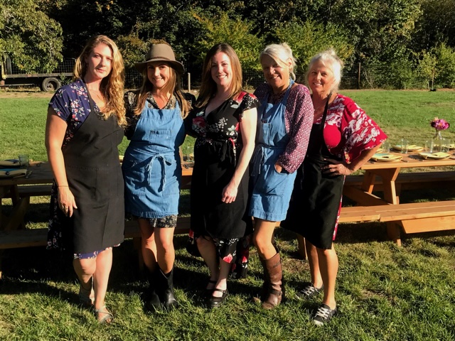 Five women standing outdoors on grass, smiling, with trees and picnic tables in the background.