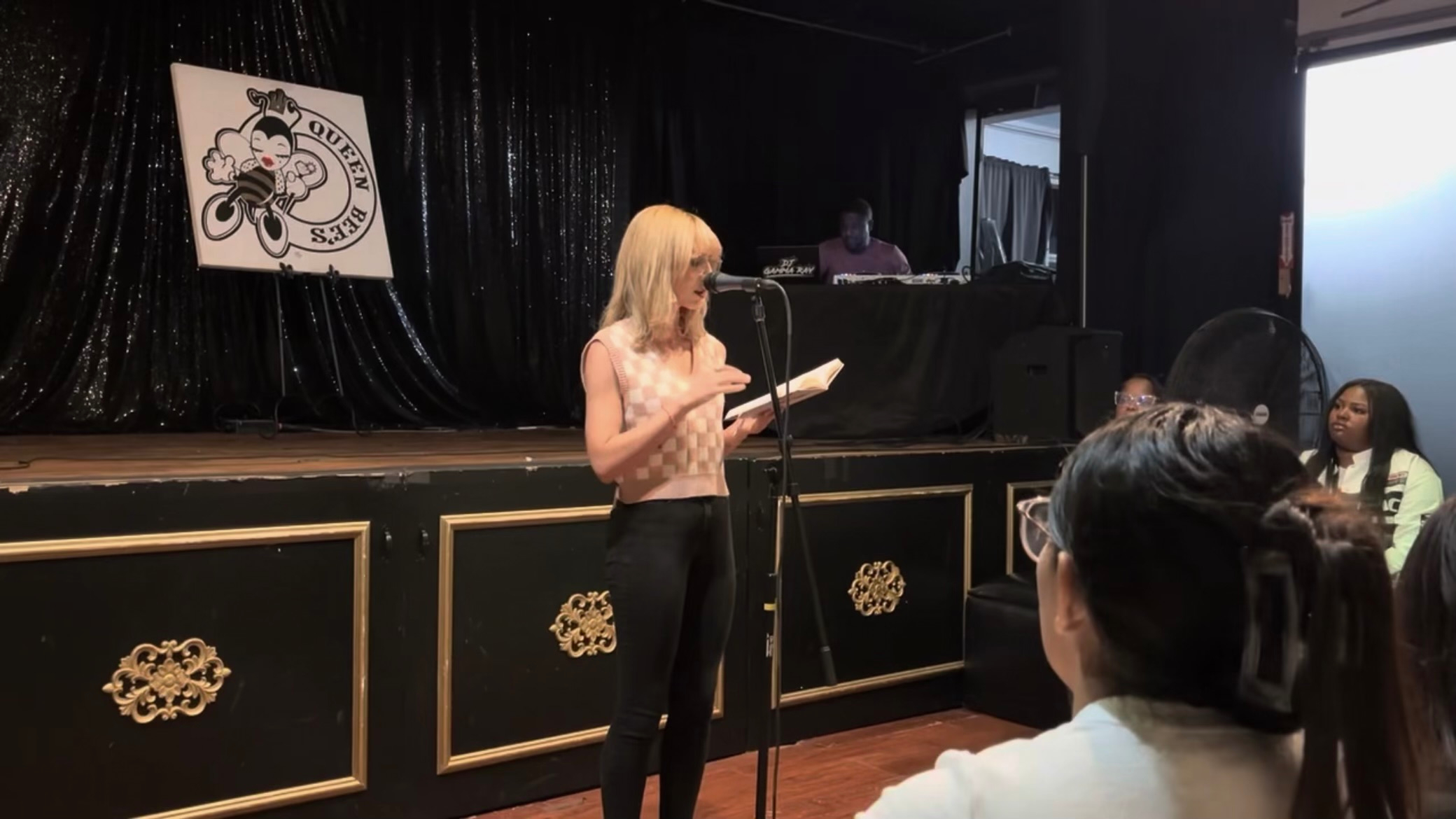 A woman with blonde hair reads from a paper on stage, with an audience watching, in a room with black curtains.