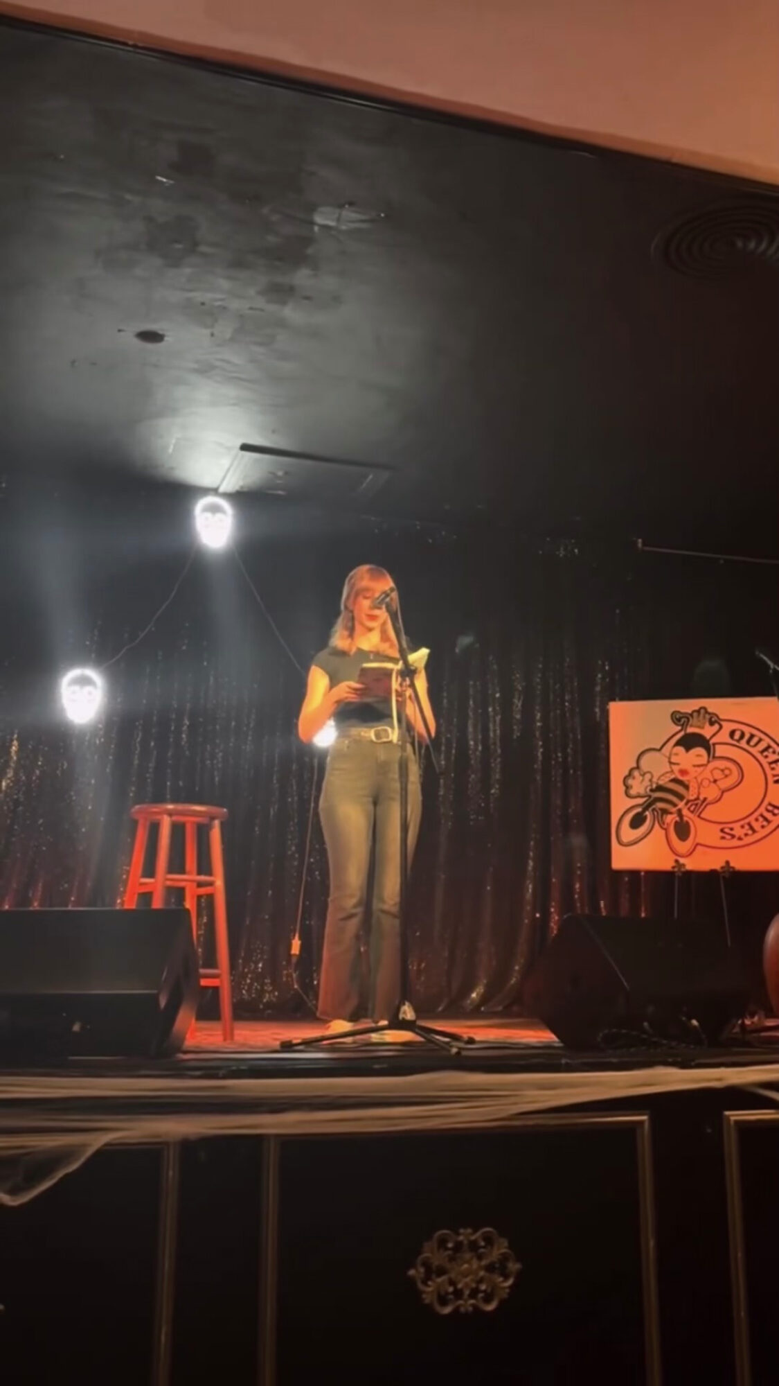 Person standing on stage with a microphone, black curtain background, orange stool, and a sign with a motorcycle illustration.
