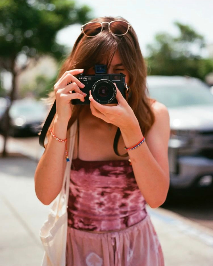 Person taking a photo outdoors, wearing sunglasses on head, pink top, and bracelets, with cars and trees in background.