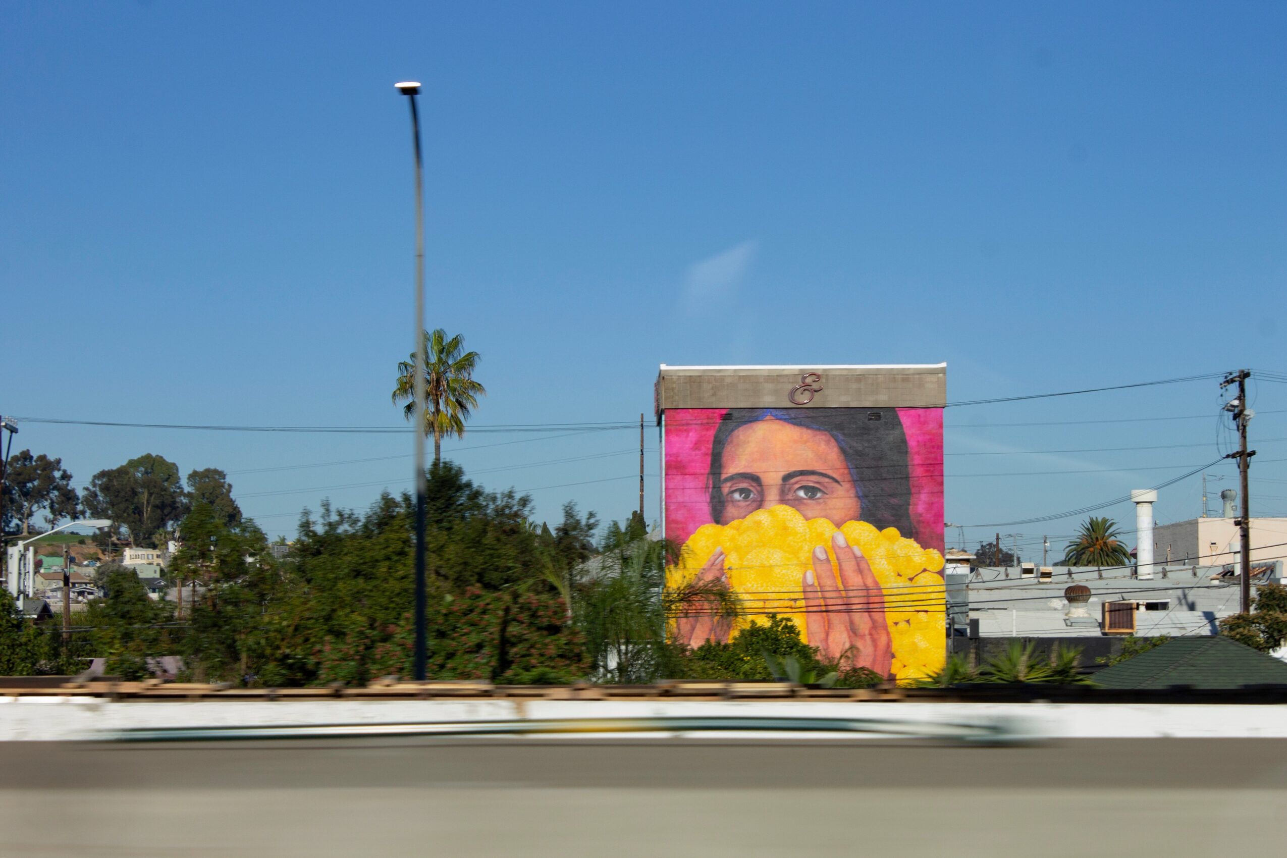 Building with a mural of a woman holding a yellow object, pink background, trees, and a clear blue sky.