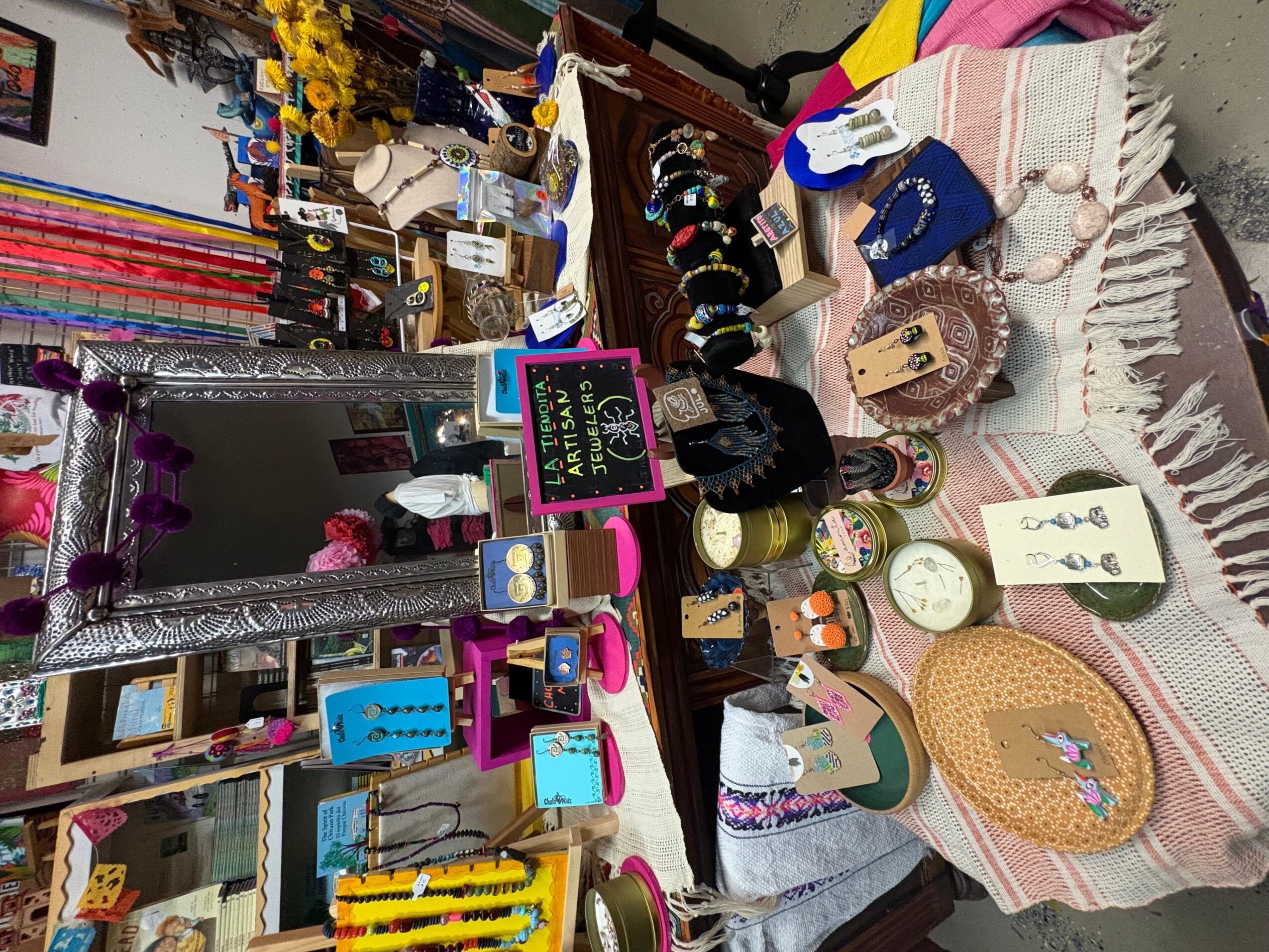 Display of jewelry, accessories, and decorative items on tables and shelves, with a mirror and colorful signs, in a boutique.