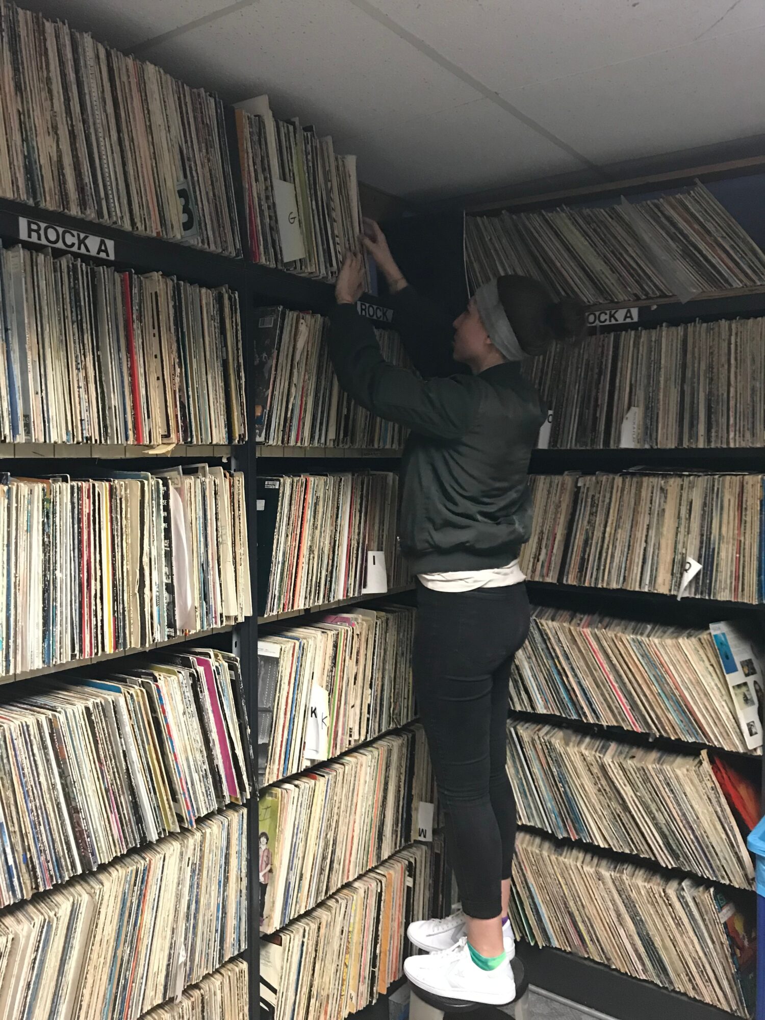 Person reaching for records on high shelf in a vinyl collection room, surrounded by shelves filled with records.