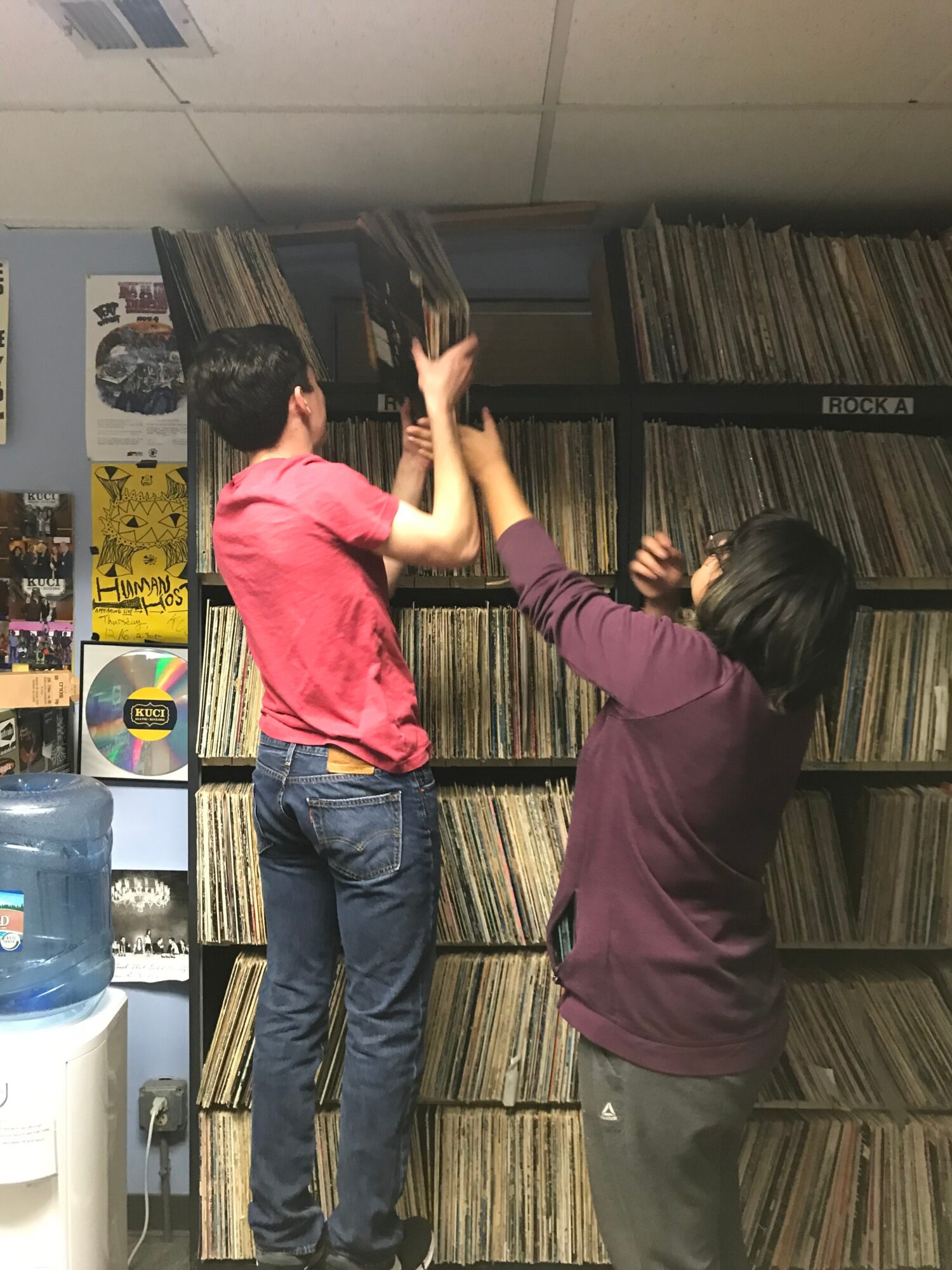 Two people reaching for records on a shelf in a record store or library, with shelves filled with vinyl records behind them.