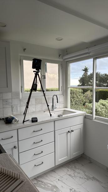 Kitchen corner with white cabinets, a window showing trees outside, and a camera on a tripod on the counter.