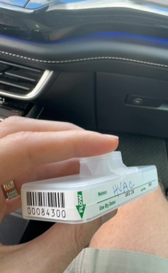 Person holding a small container with a barcode and handwritten notes inside a car, dashboard visible in background.