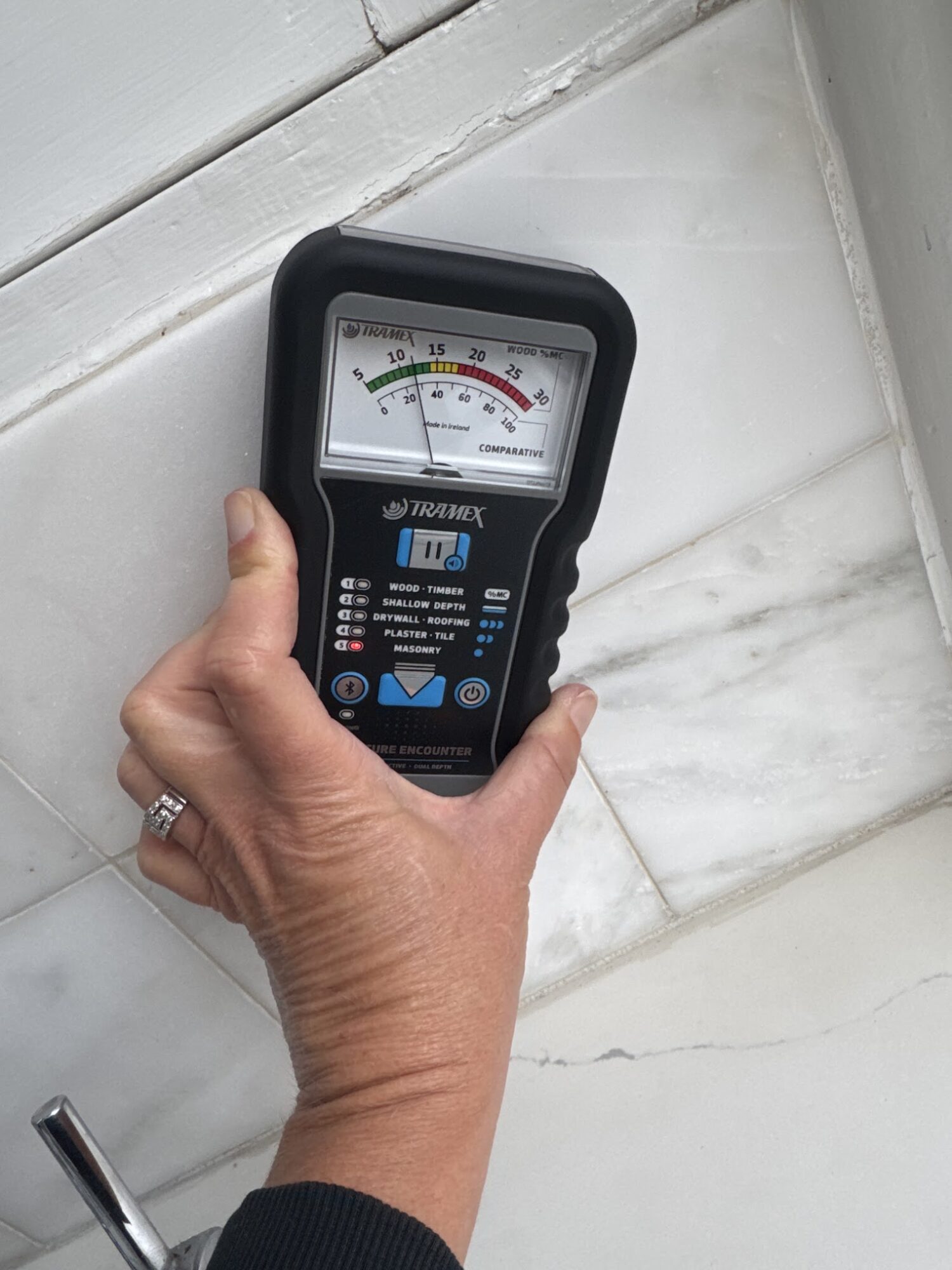 Hand holding a device with a gauge, against a white tiled wall background.