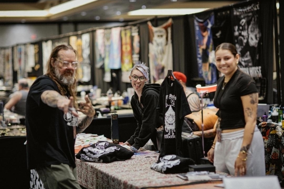 Three people at a booth with clothing and artwork displayed behind them, smiling and interacting, in an indoor market setting.