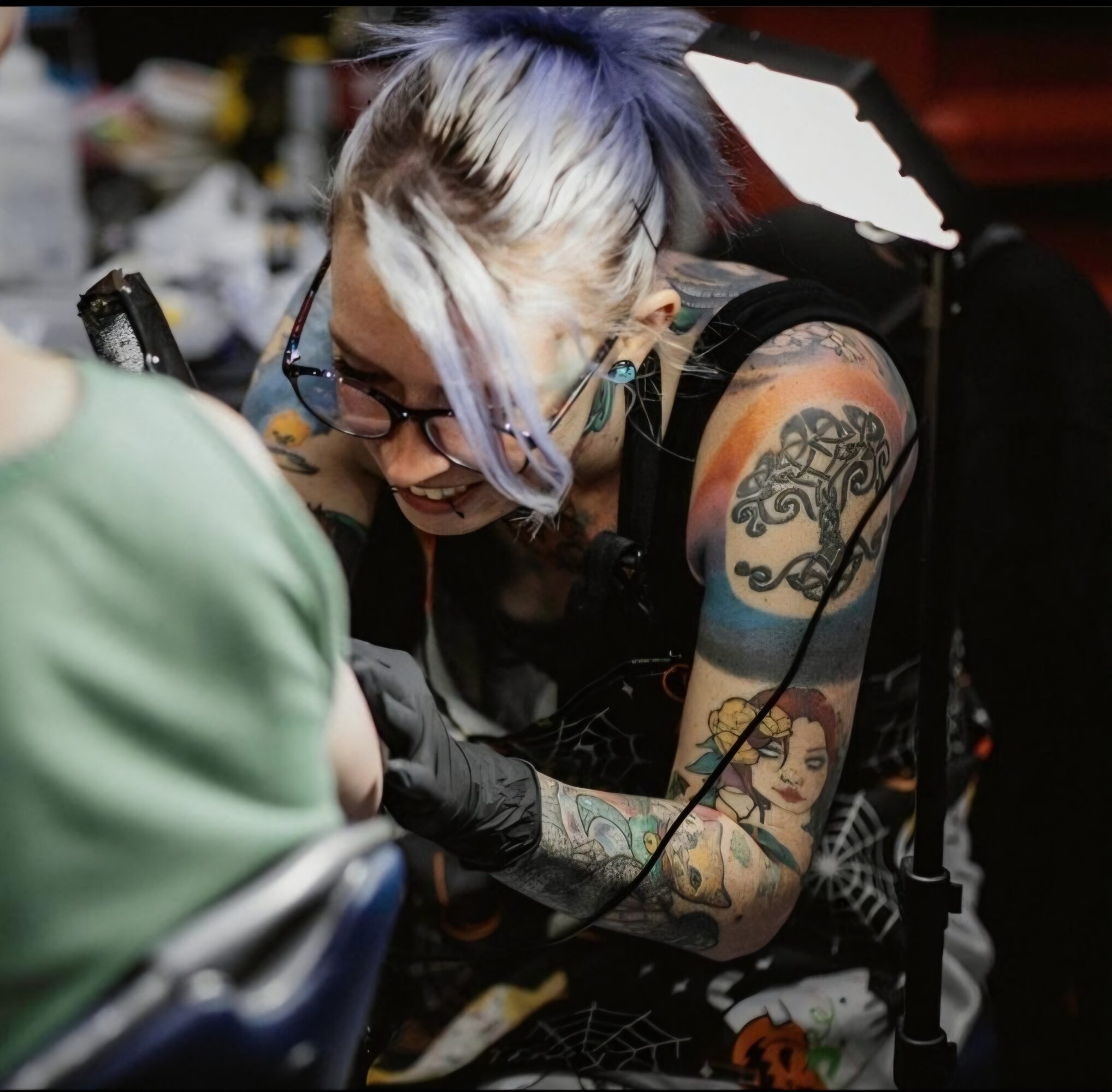 Tattooed woman with glasses and colorful hair getting a tattoo, smiling, in a tattoo studio.